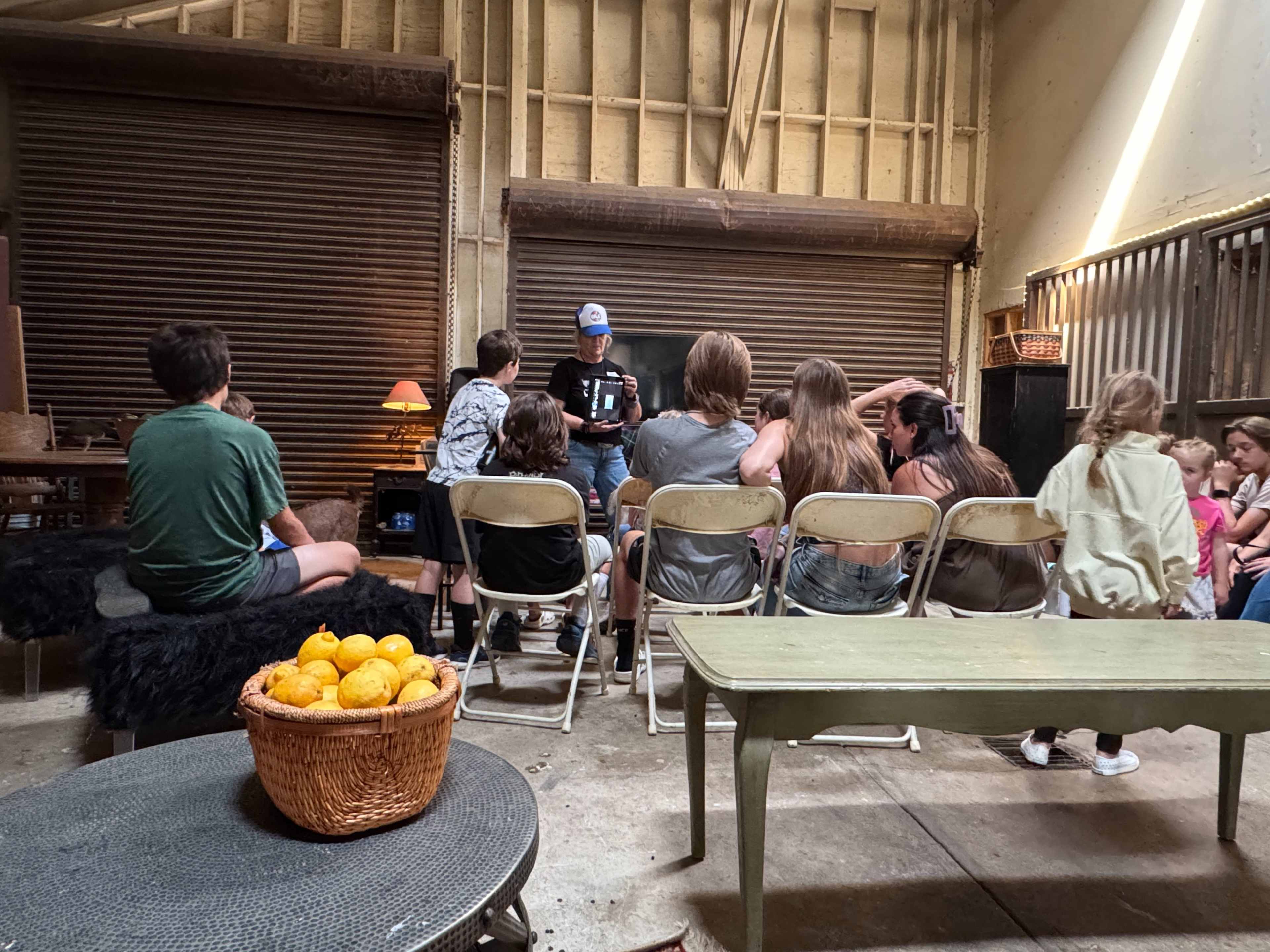 A group of children and adults are seated on folding chairs in a rustic interior, where a person is presenting to them while holding a device.