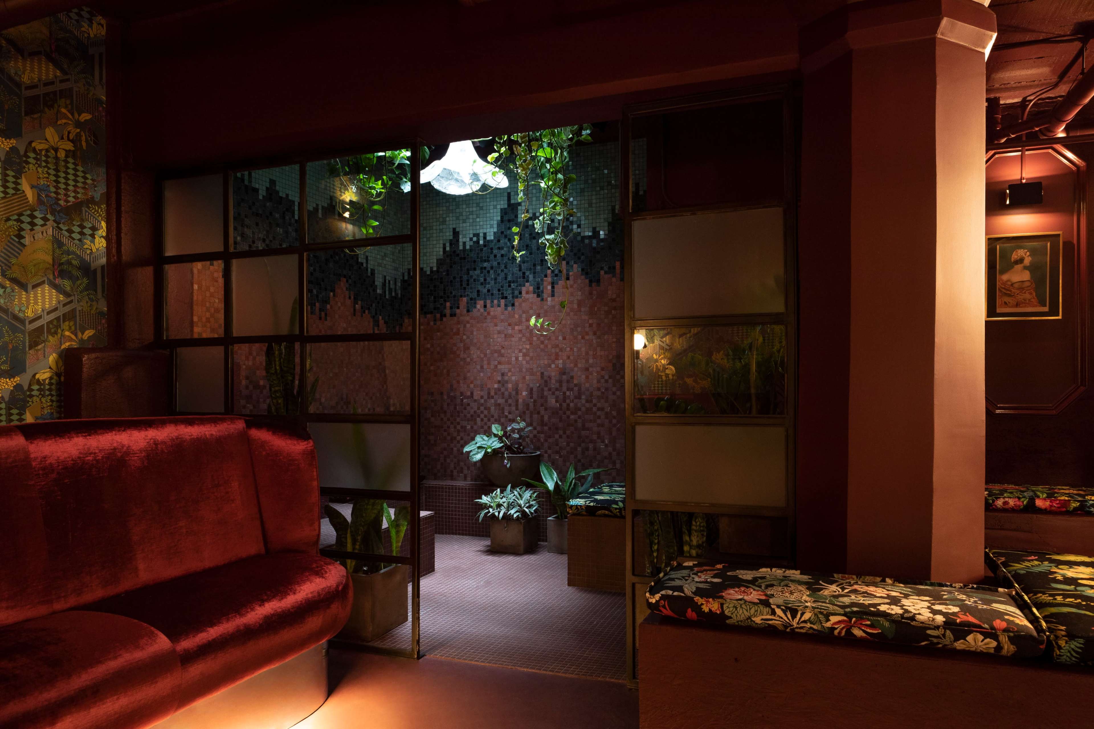 The image shows a dimly lit interior space with rich colors, featuring a seating area with a velvet couch and a view of a plant-filled room beyond a glass partition.
