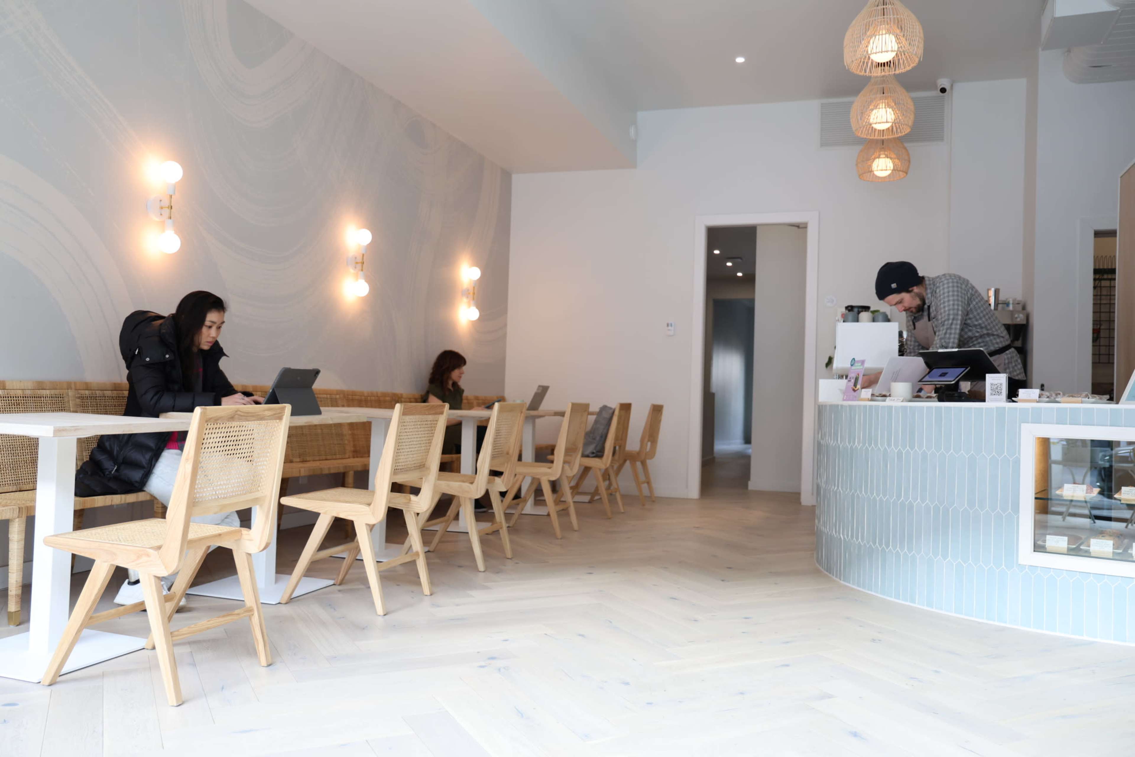 A modern café interior with light wooden flooring, a counter at the back, and two women working on laptops at tables.