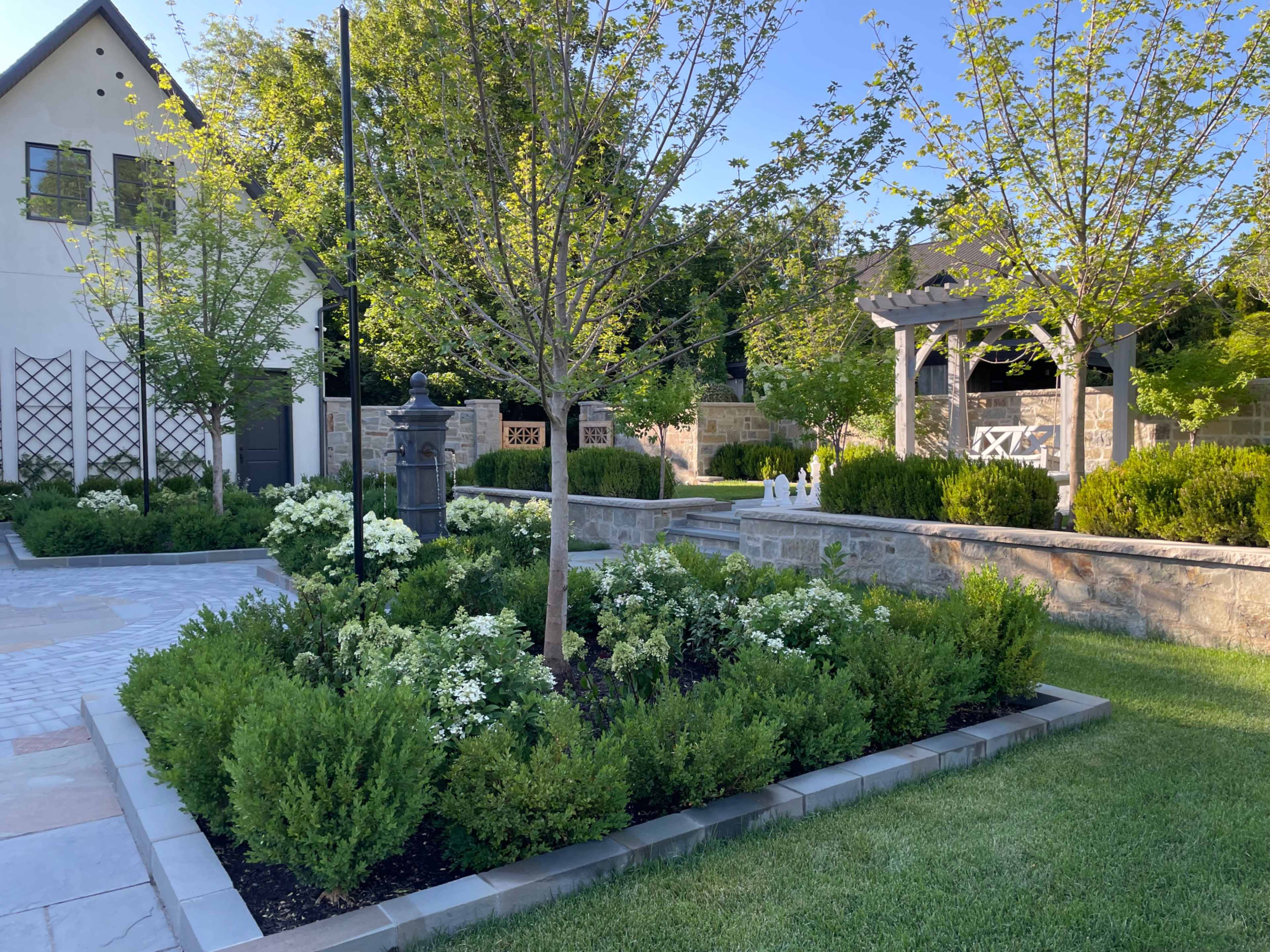 A landscaped garden features neatly arranged flower beds, young trees, and a stone pathway leading to a gazebo and stone walls.