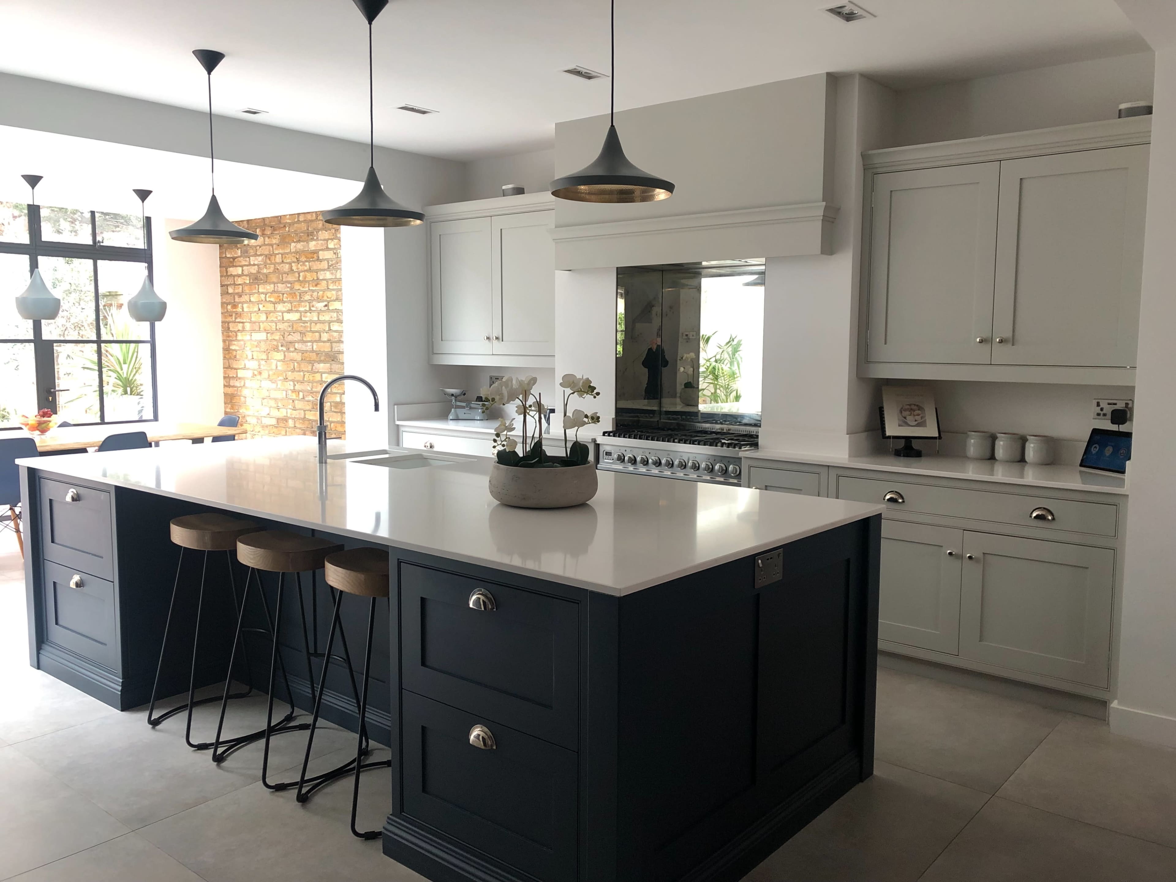 The image shows a modern kitchen featuring a central island with bar stools, light-colored cabinets, dark blue accents, and large windows letting in natural light.