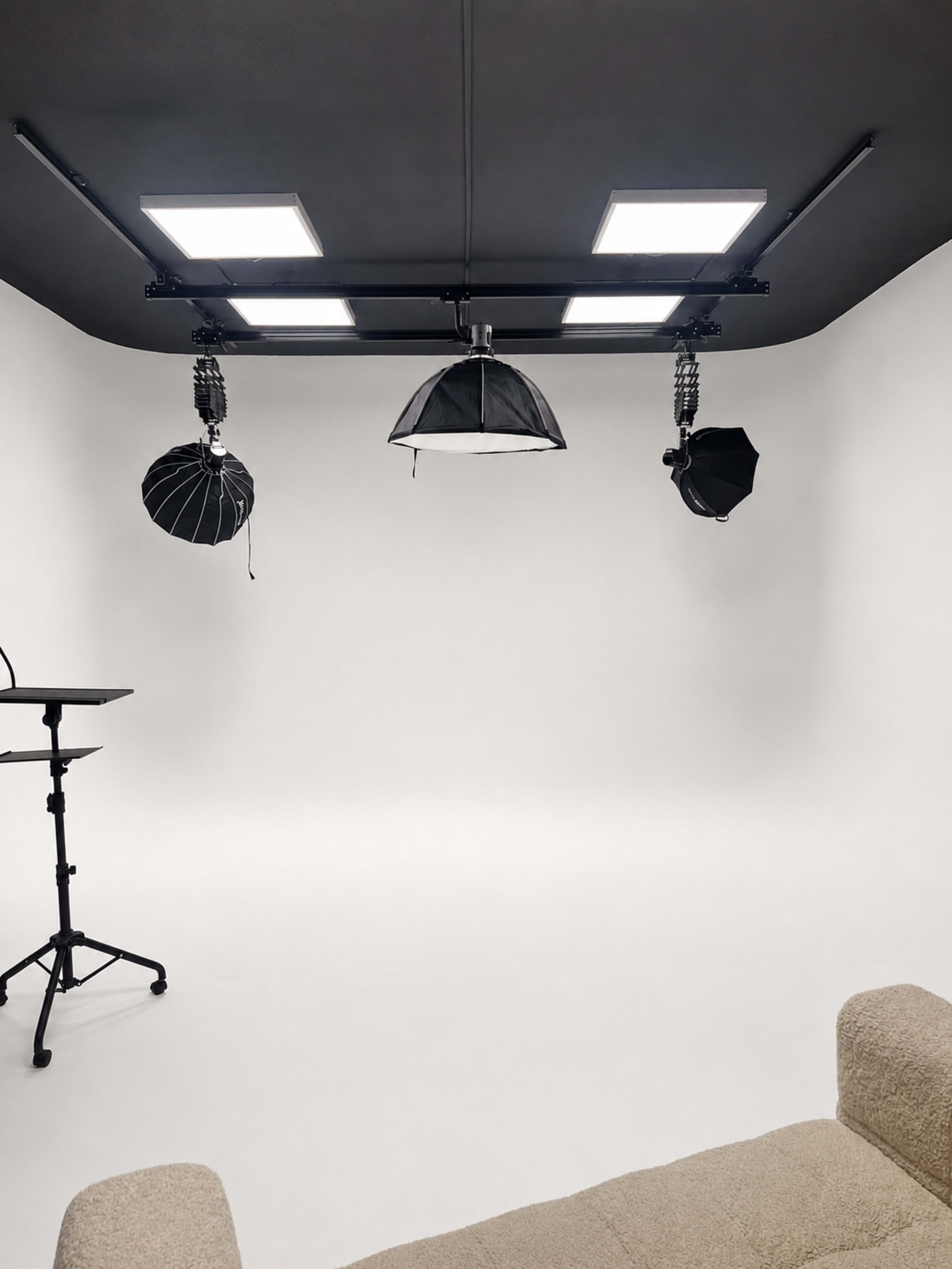 The image shows a minimalist photo studio with a white background, equipped with four lighting fixtures and a beige sofa.