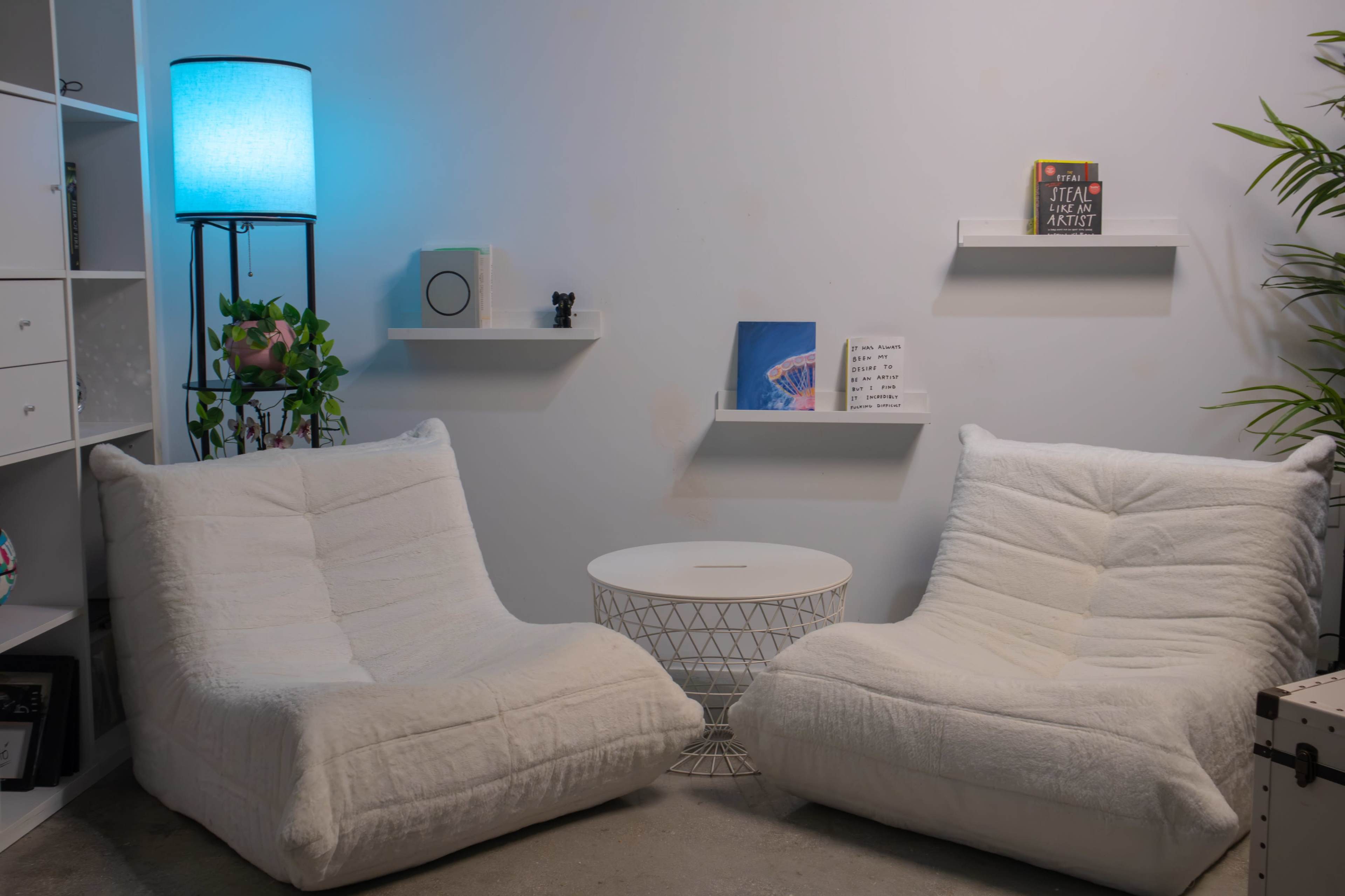The image shows a cozy seating area with two white, plush chairs, a round white table, and decorative shelves on a light-colored wall.