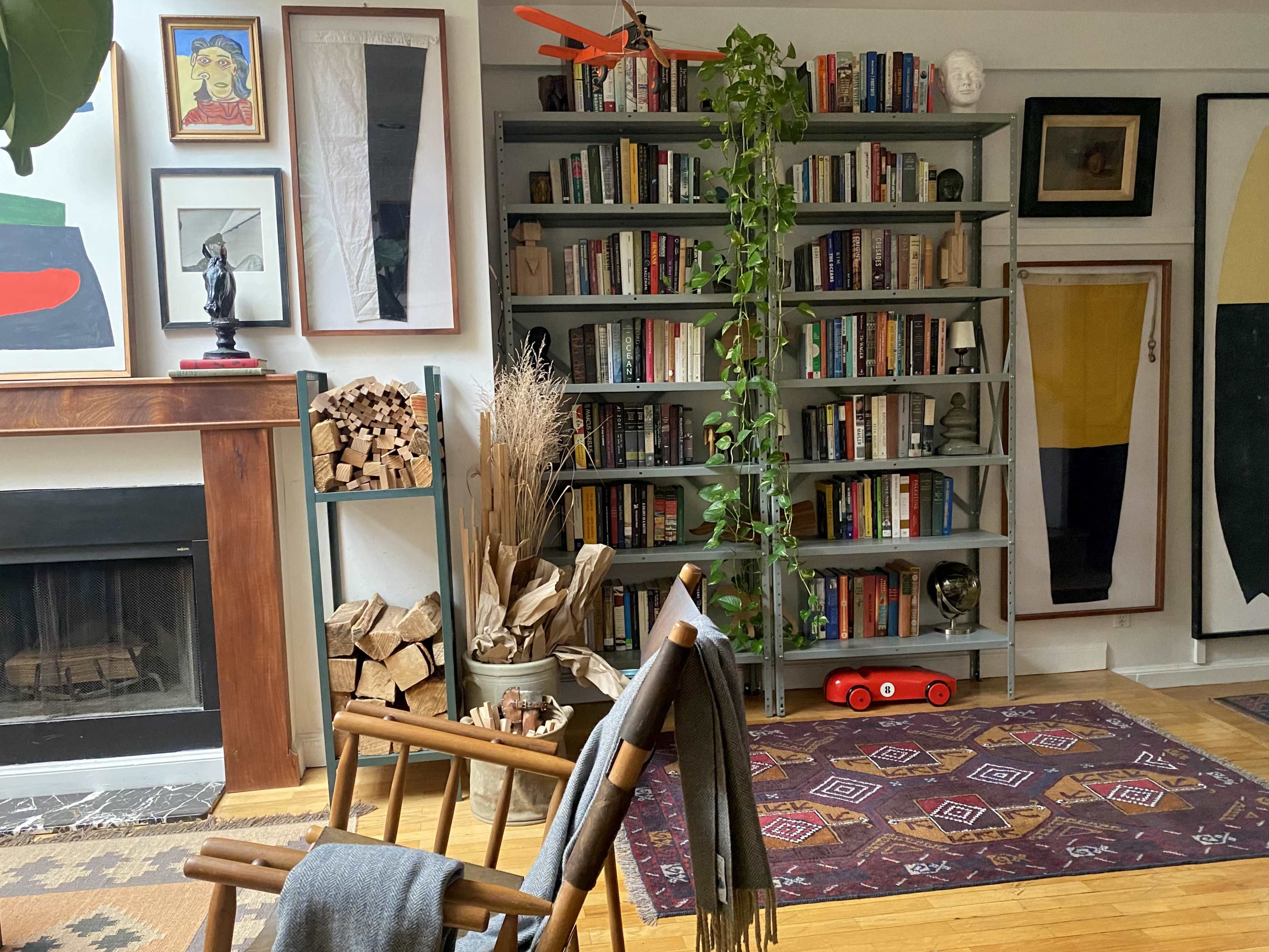 The image shows a cozy interior with a bookshelf filled with books, a fireplace, a stack of firewood, and a red toy car on a wooden floor.