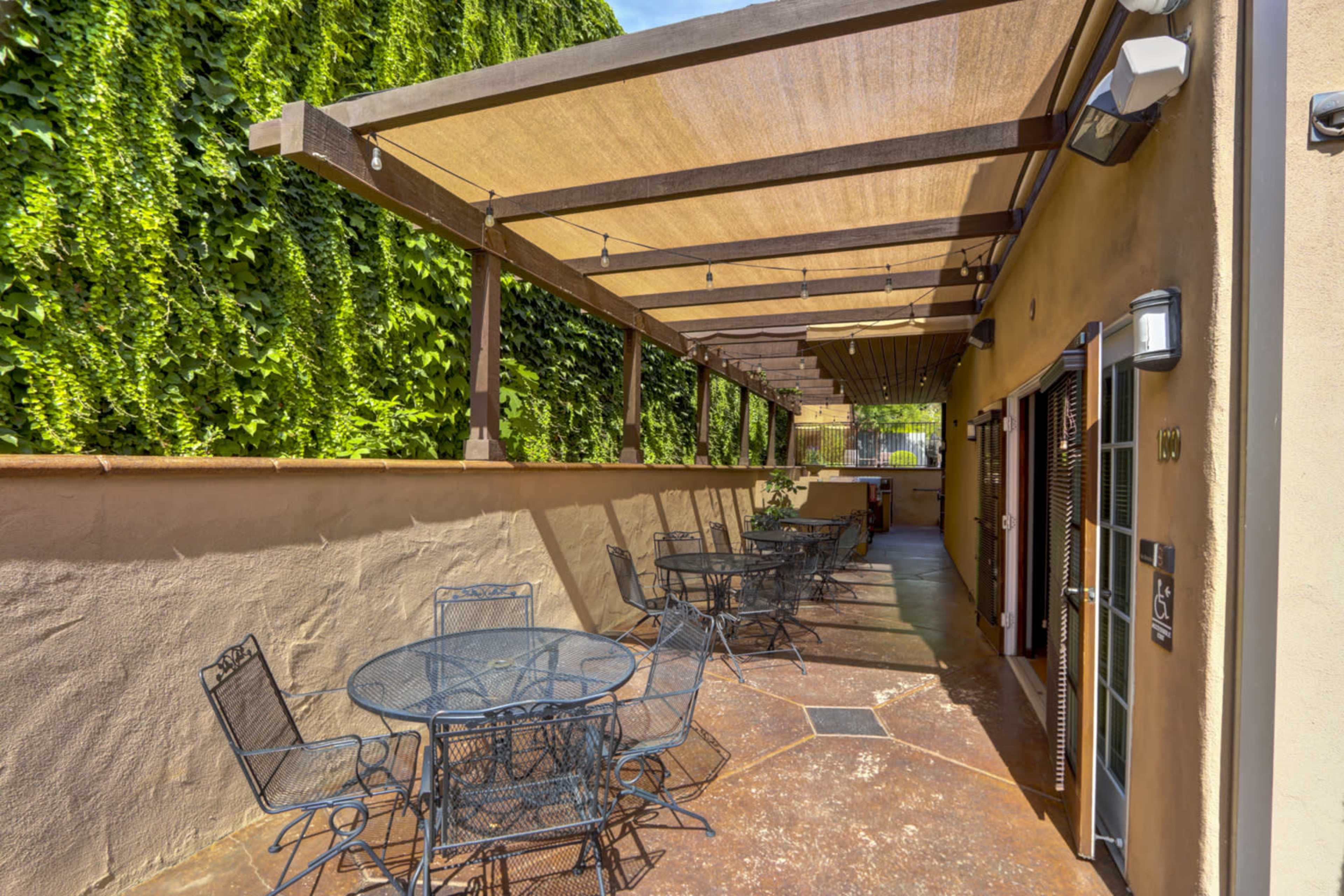 A patio area with several round tables and chairs shaded by a large canopy and surrounded by green foliage.