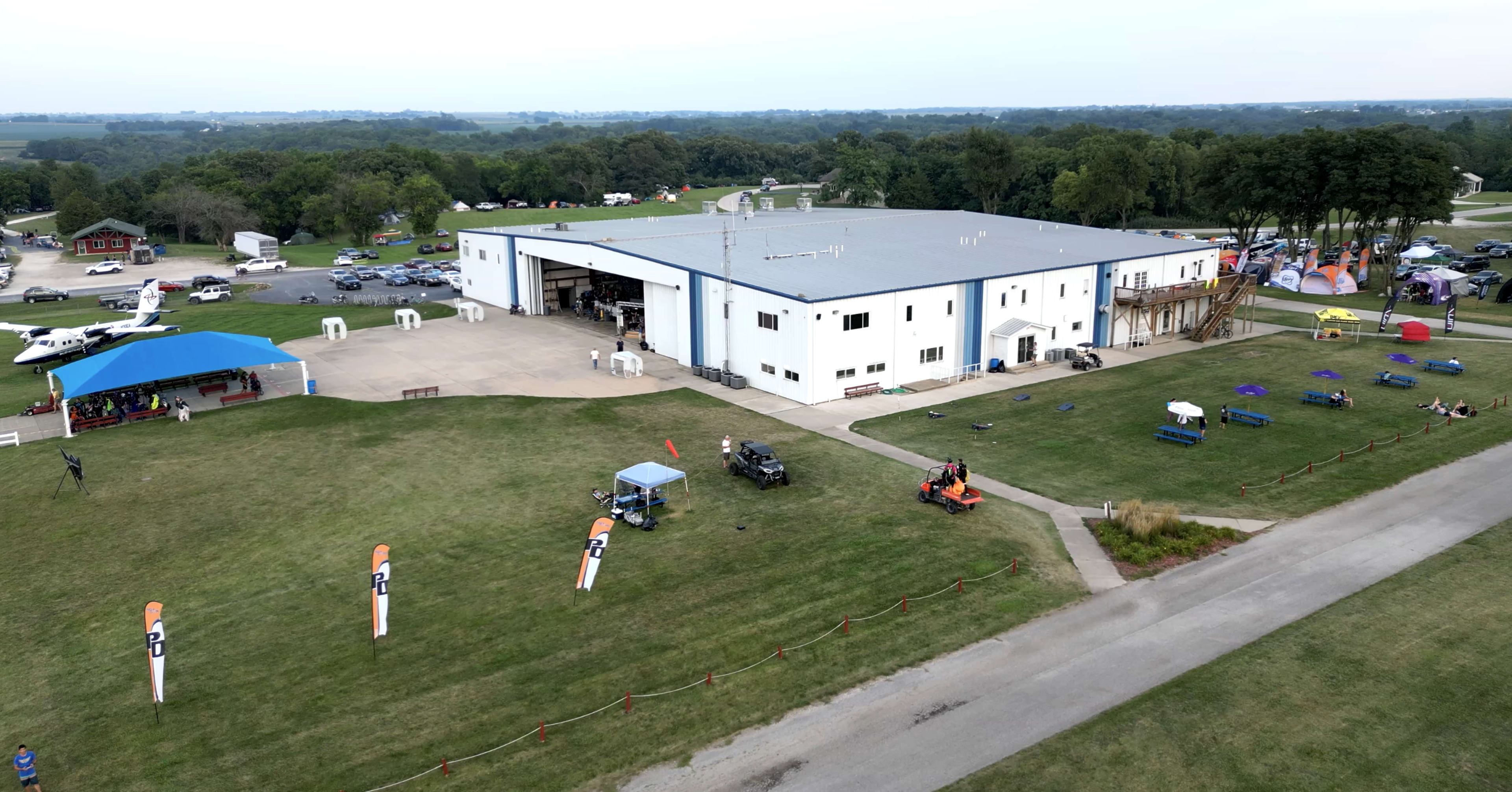 The image shows a large white building with a gray roof, surrounded by grassy areas, parking lots, and various vehicles parked nearby.