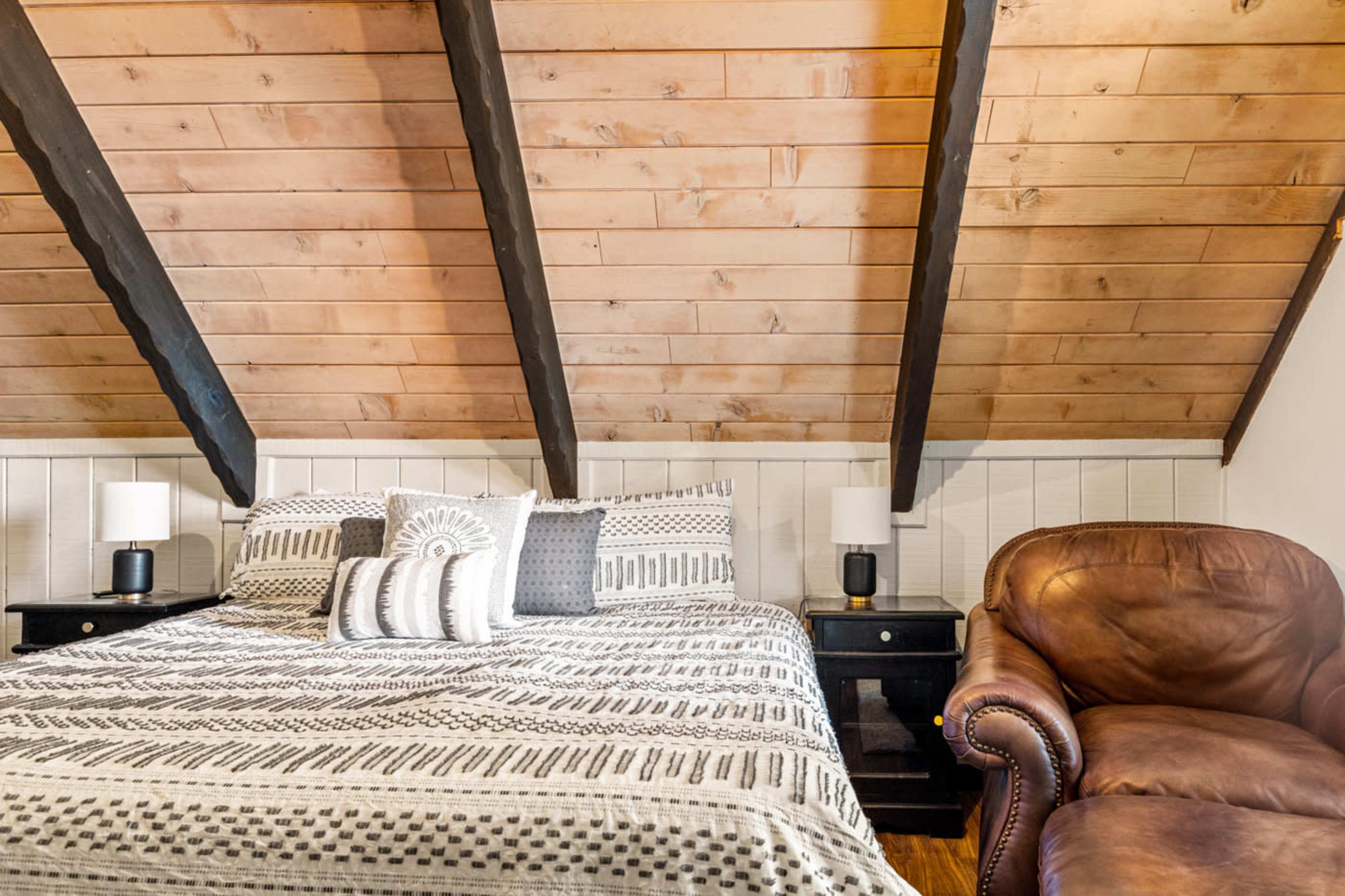 The image shows a cozy attic bedroom featuring a bed with patterned bedding, two bedside lamps, and a leather armchair against a wall with wooden beams.