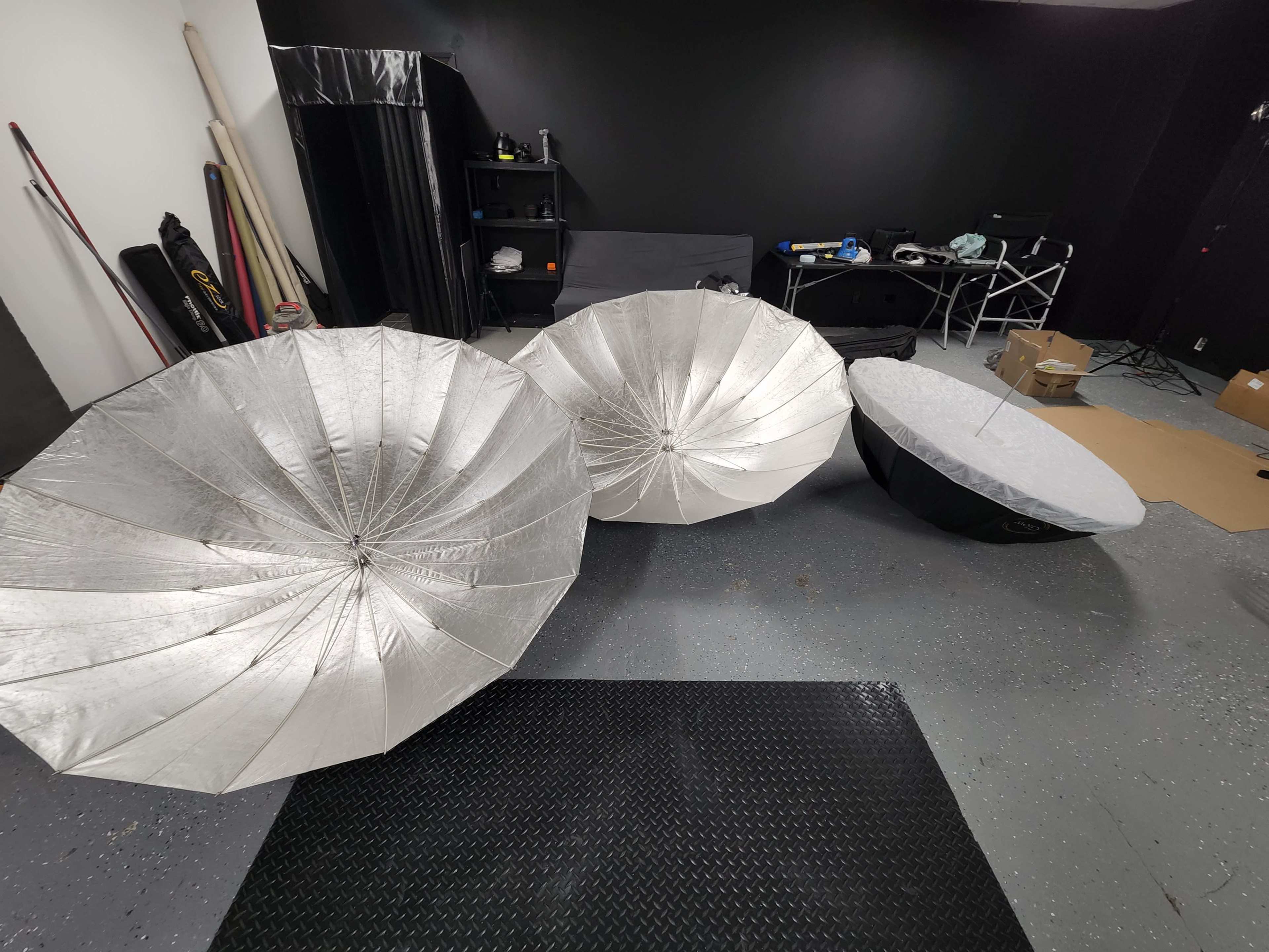 Three large reflective umbrella softboxes are set up on a gray floor in a studio space with various equipment and furniture in the background.