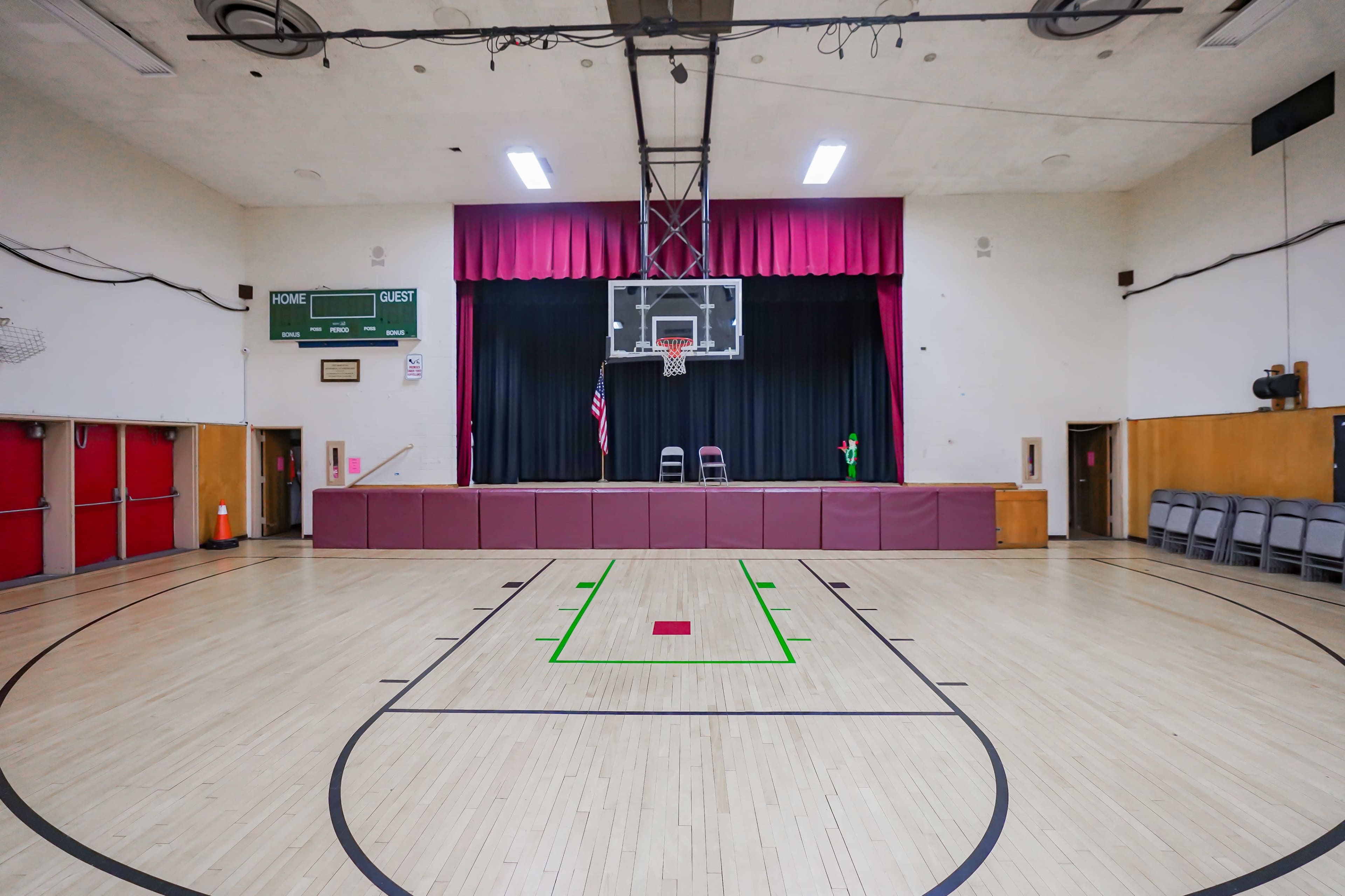 The image shows an empty gymnasium with a basketball court, a stage at the back, and a scoreboard on the wall.