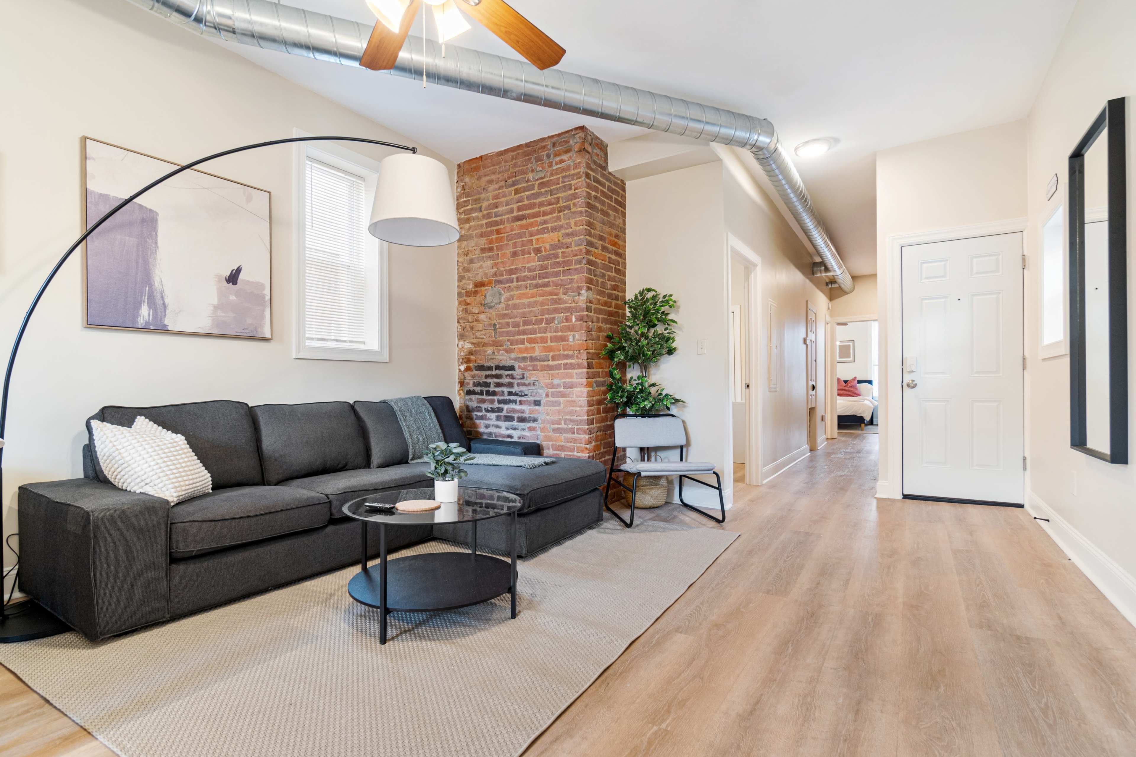 The image features a living room with a dark gray sofa, a round black coffee table, and exposed brick walls, leading to a hallway with doors on either side.