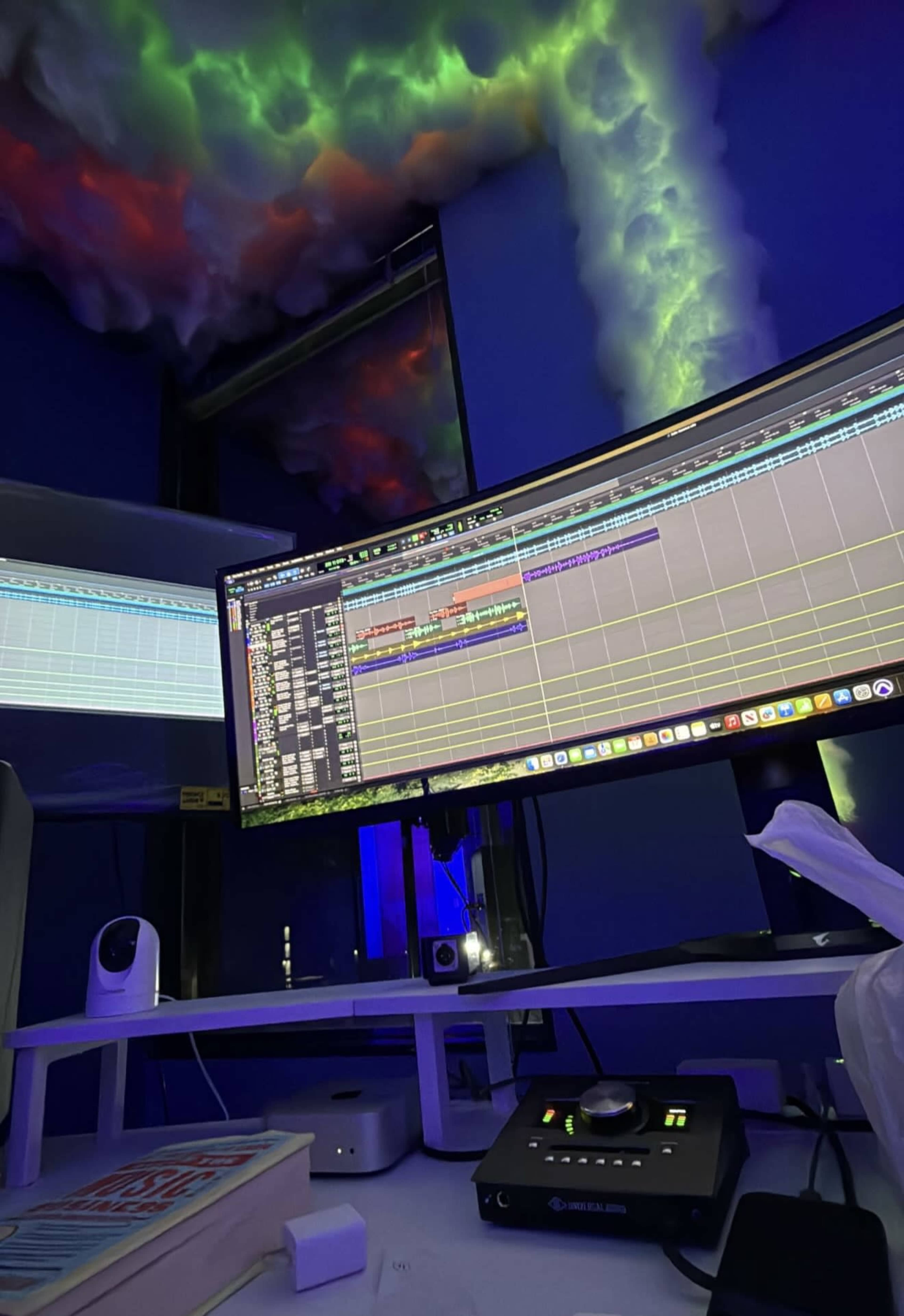 A computer setup features two monitors displaying digital audio editing software in a dimly lit room with colorful ambient lighting.