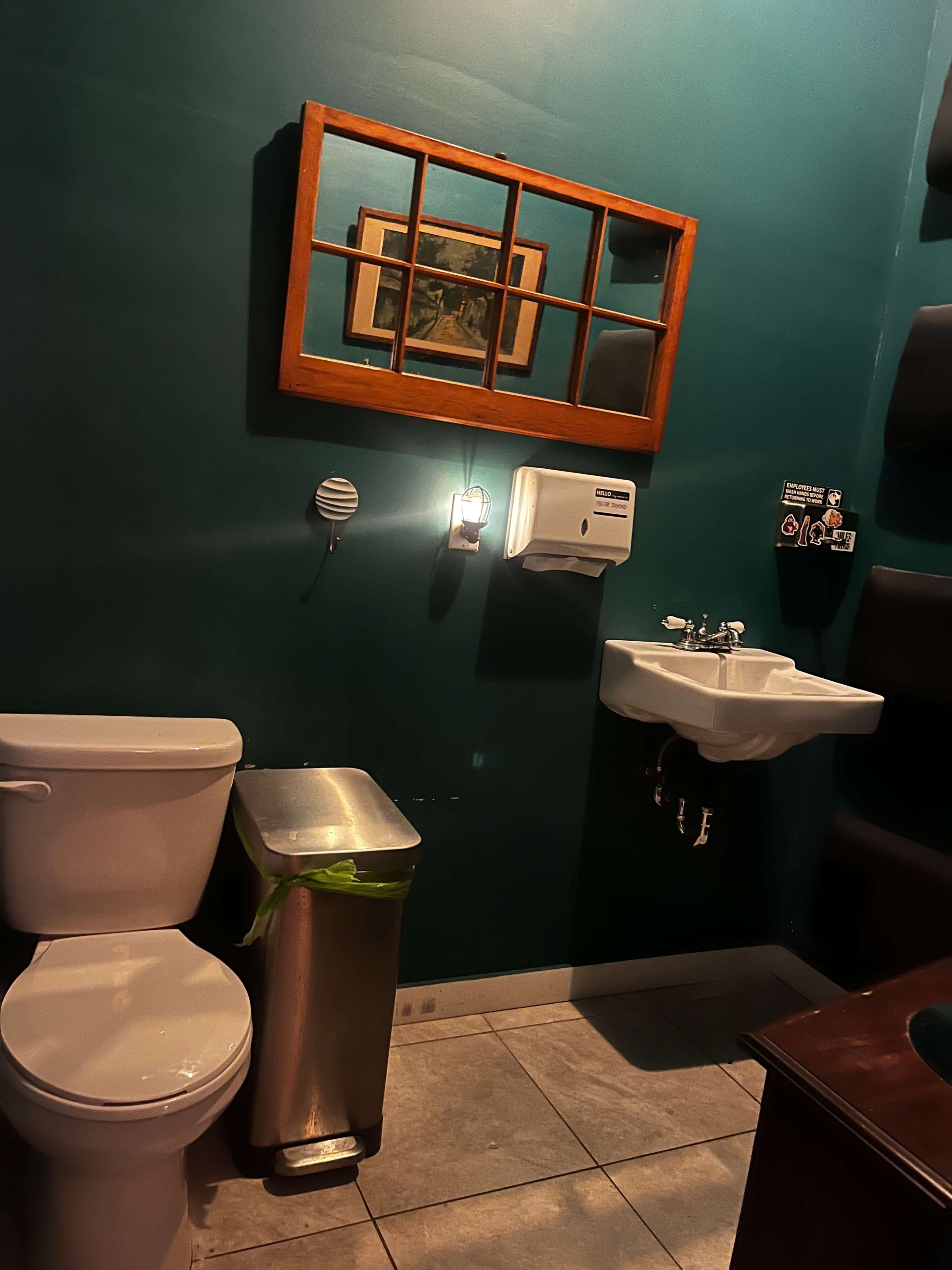 A small bathroom featuring a toilet, a sink, a mirror framed in wood, a paper towel dispenser, and a trash can.