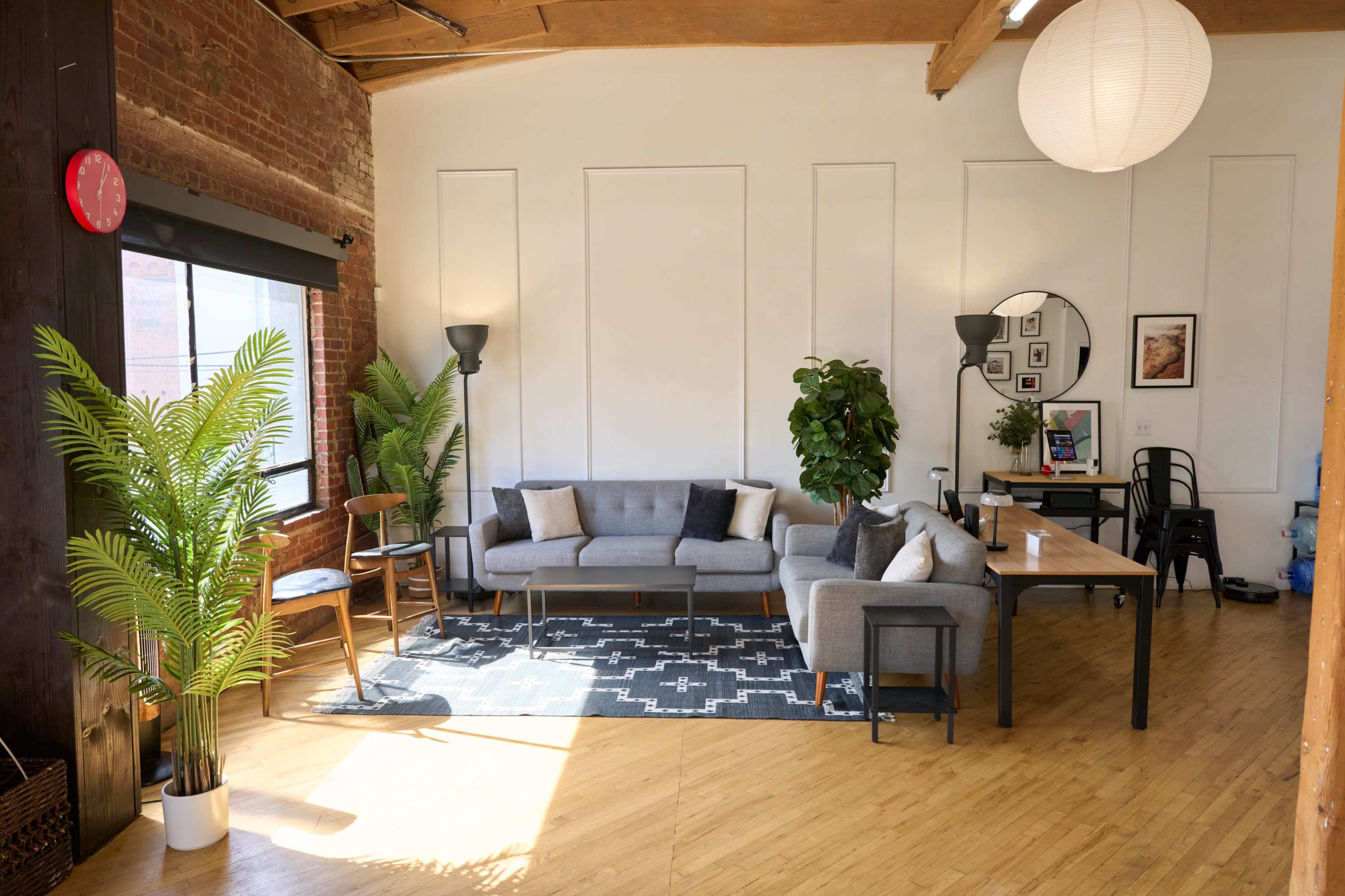 The image shows a bright and spacious living area featuring a gray sectional sofa, a wooden dining table, indoor plants, and decorative wall art.