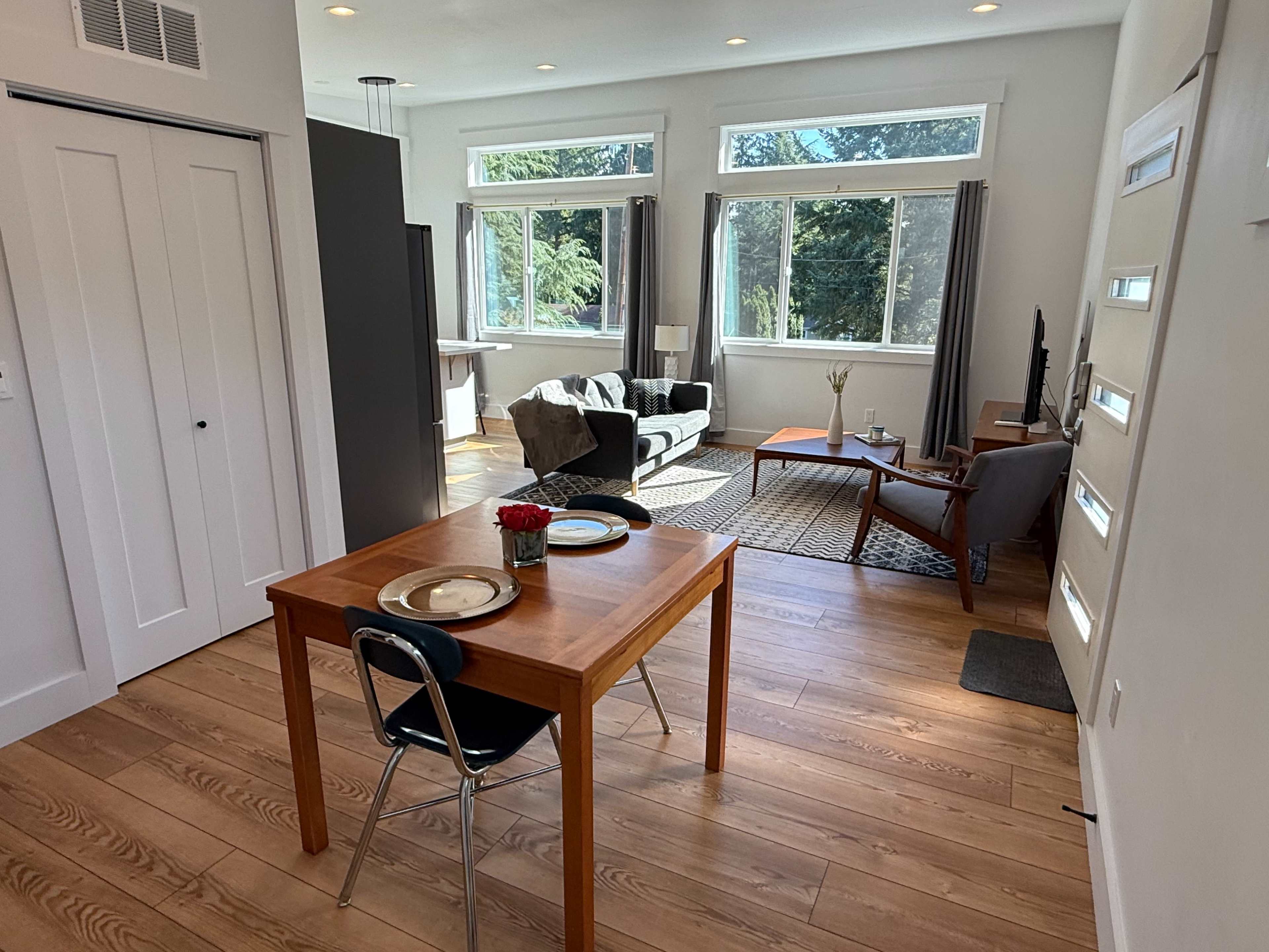 A small dining area with a wooden table and chairs opens up to a cozy living space featuring a sofa and armchair, illuminated by natural light through large windows.