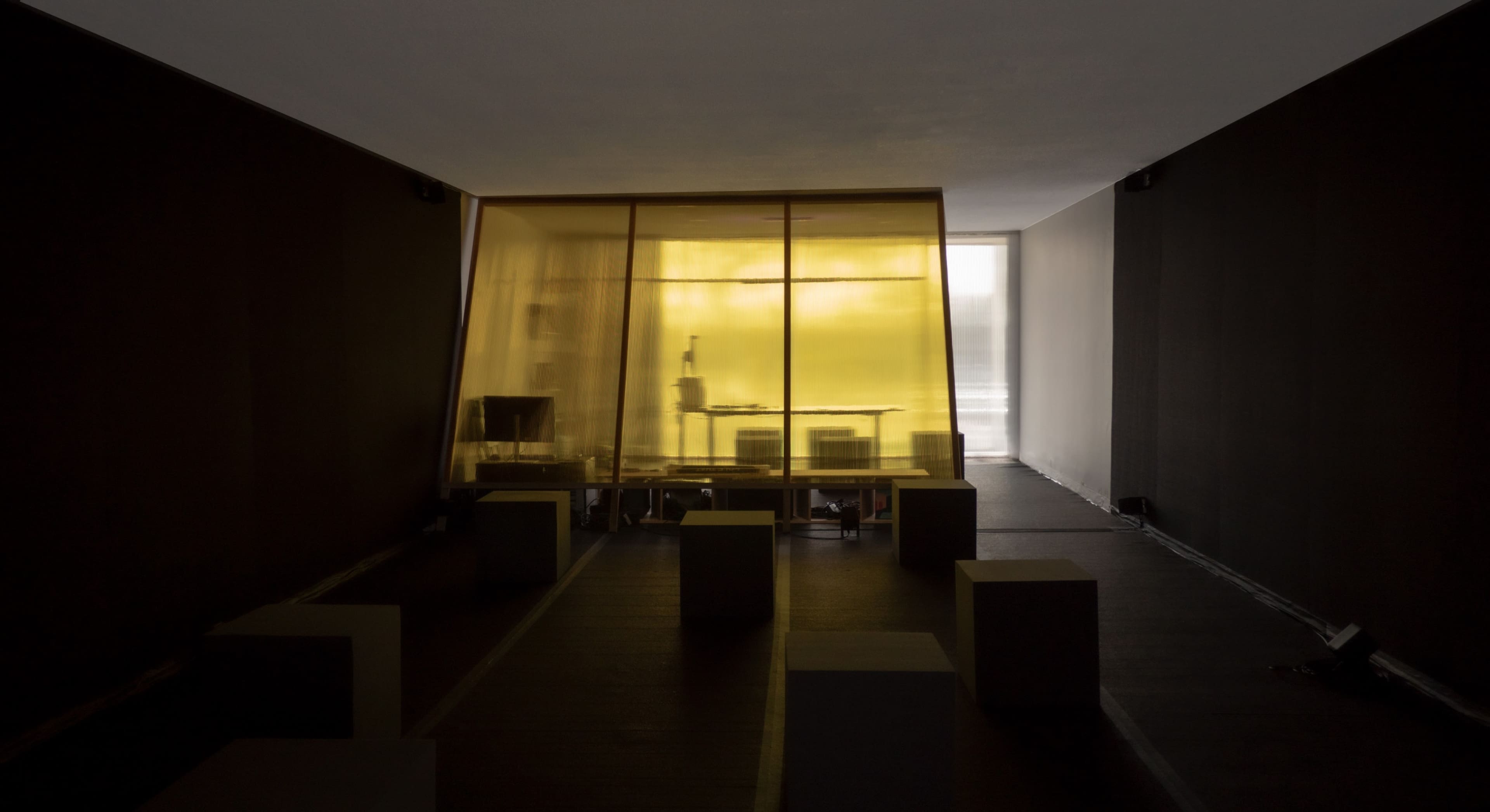 A room features a yellow-tinted glass enclosure containing a workstation, surrounded by gray, cube-shaped seating.