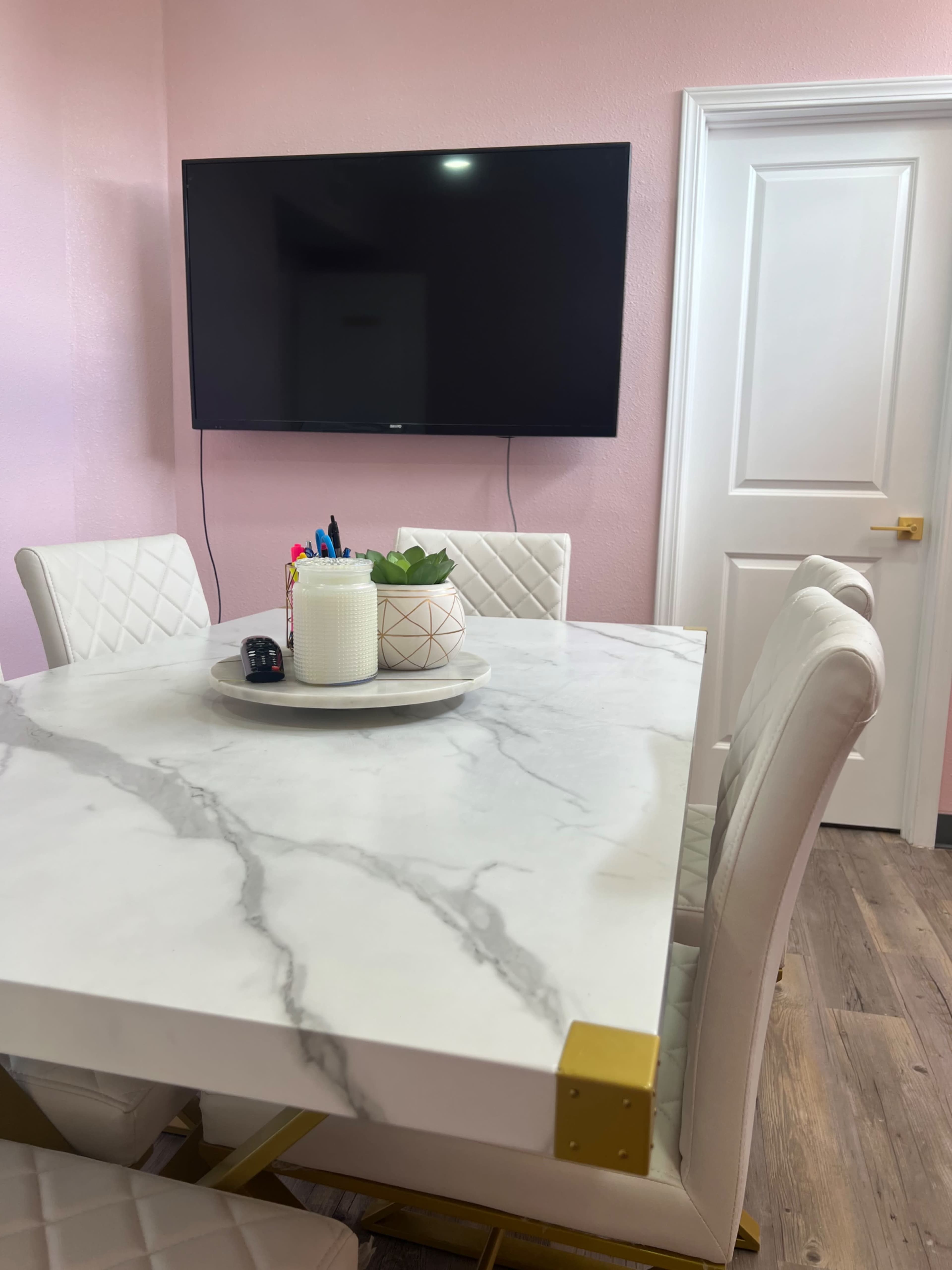 A marble dining table with a decorative centerpiece is set against a pink wall, featuring a television mounted nearby and several white upholstered chairs.