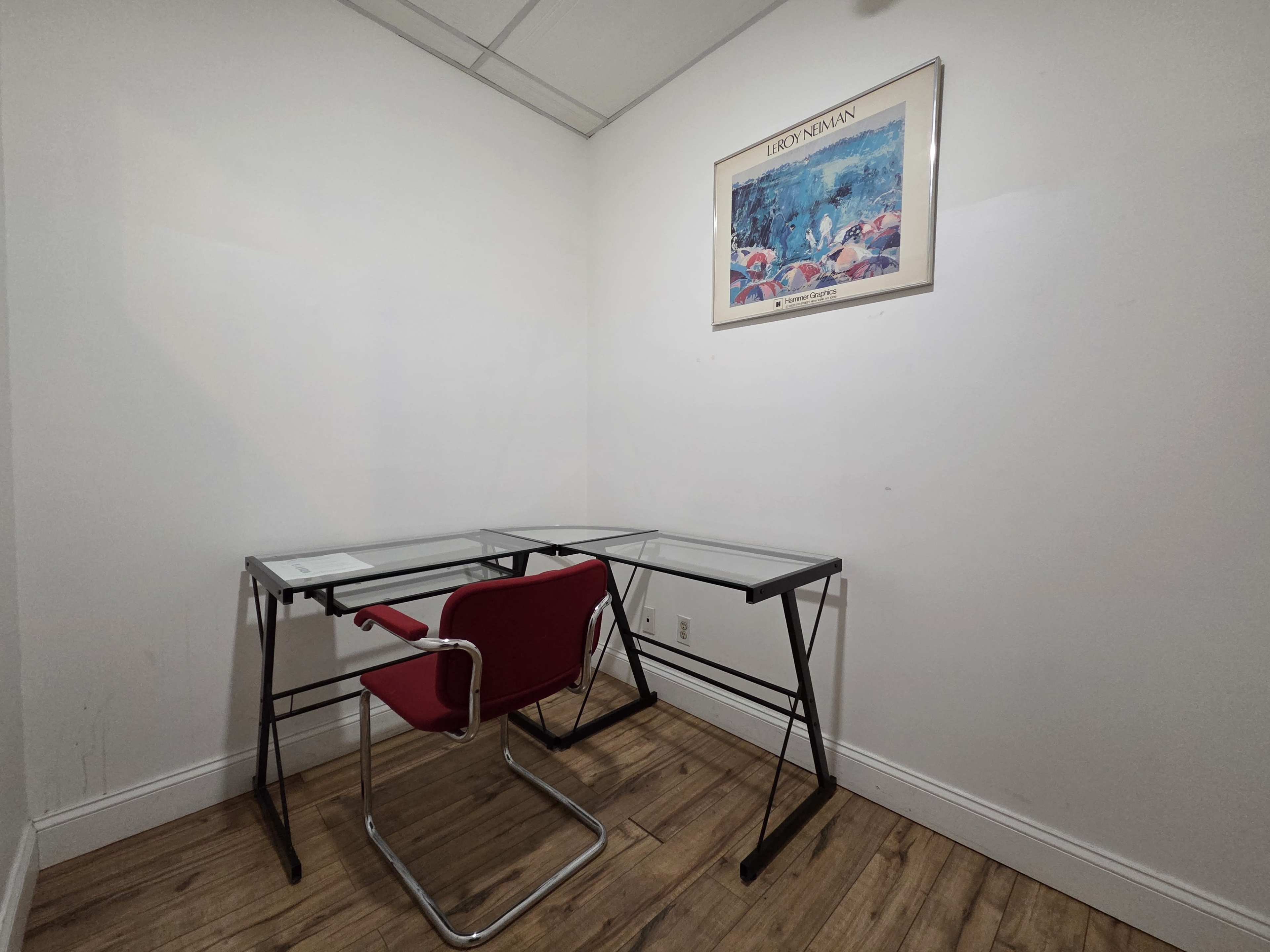 The image shows a small office space featuring a glass-topped desk, a red chair, and a framed artwork on the wall.