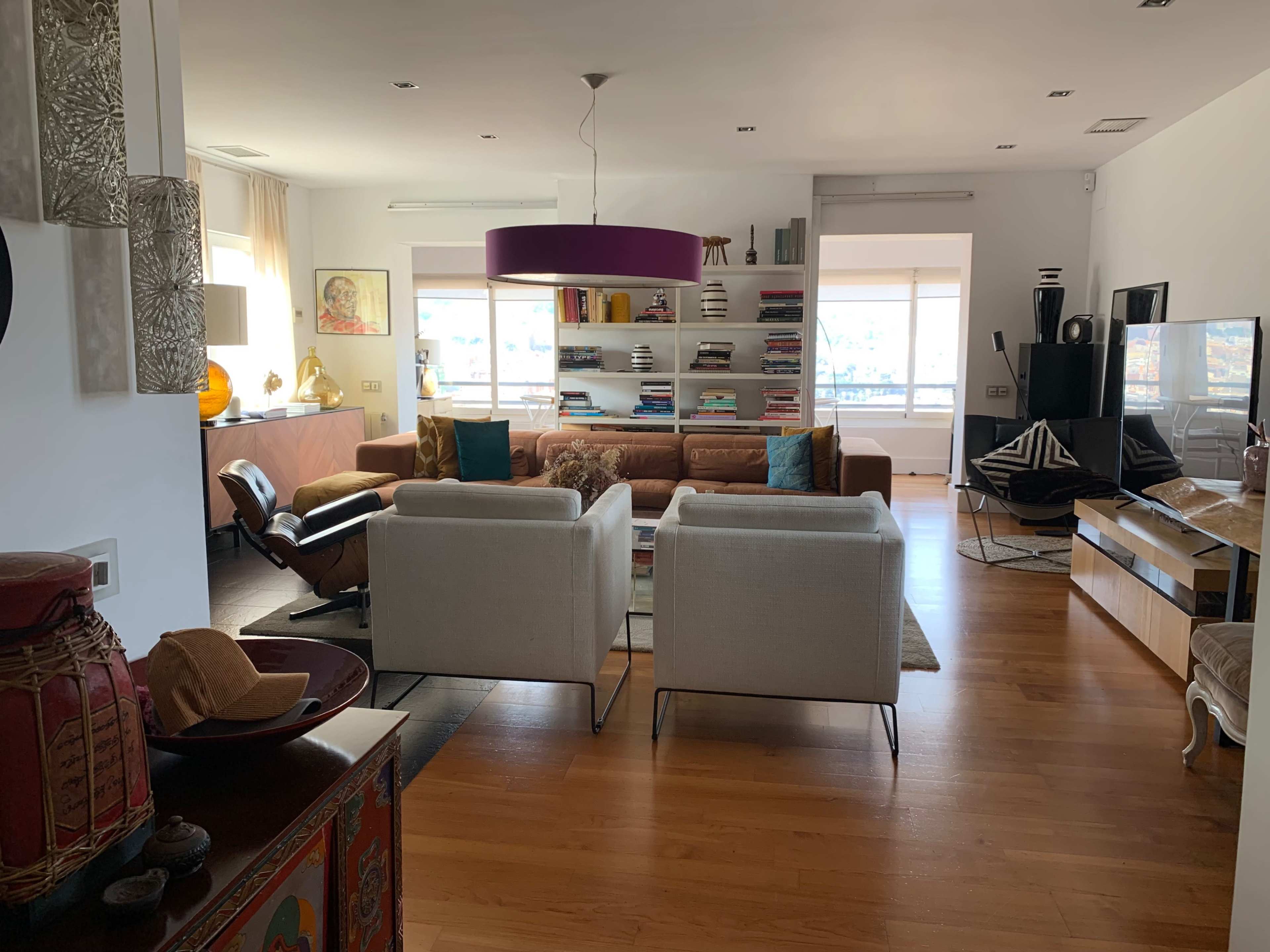 A modern living room with a sofa, two armchairs, bookshelves, and large windows allowing natural light.