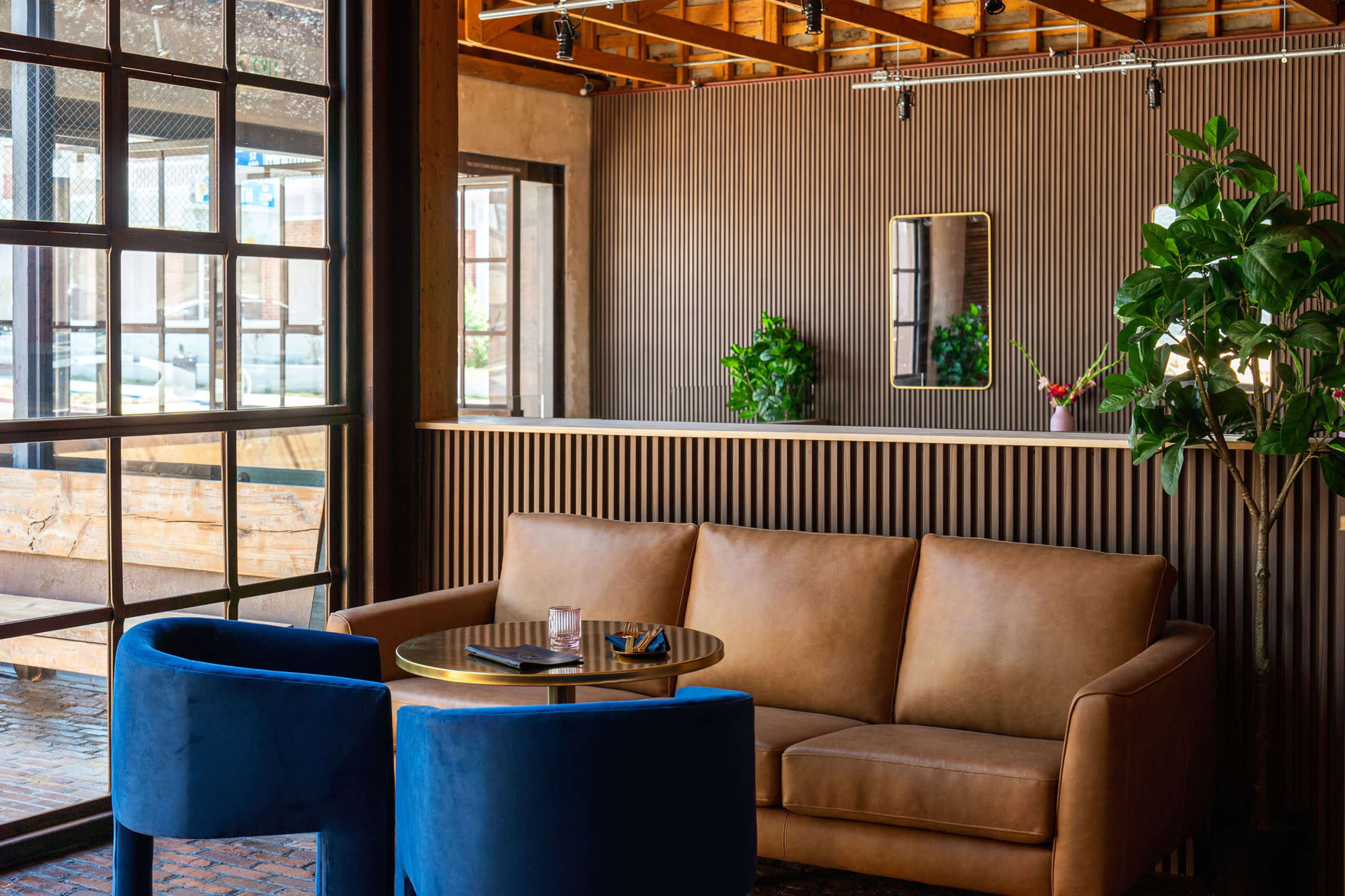 A cozy lounge area with a brown leather sofa, two blue chairs, a round table, and a wall featuring a mirror and potted plants.
