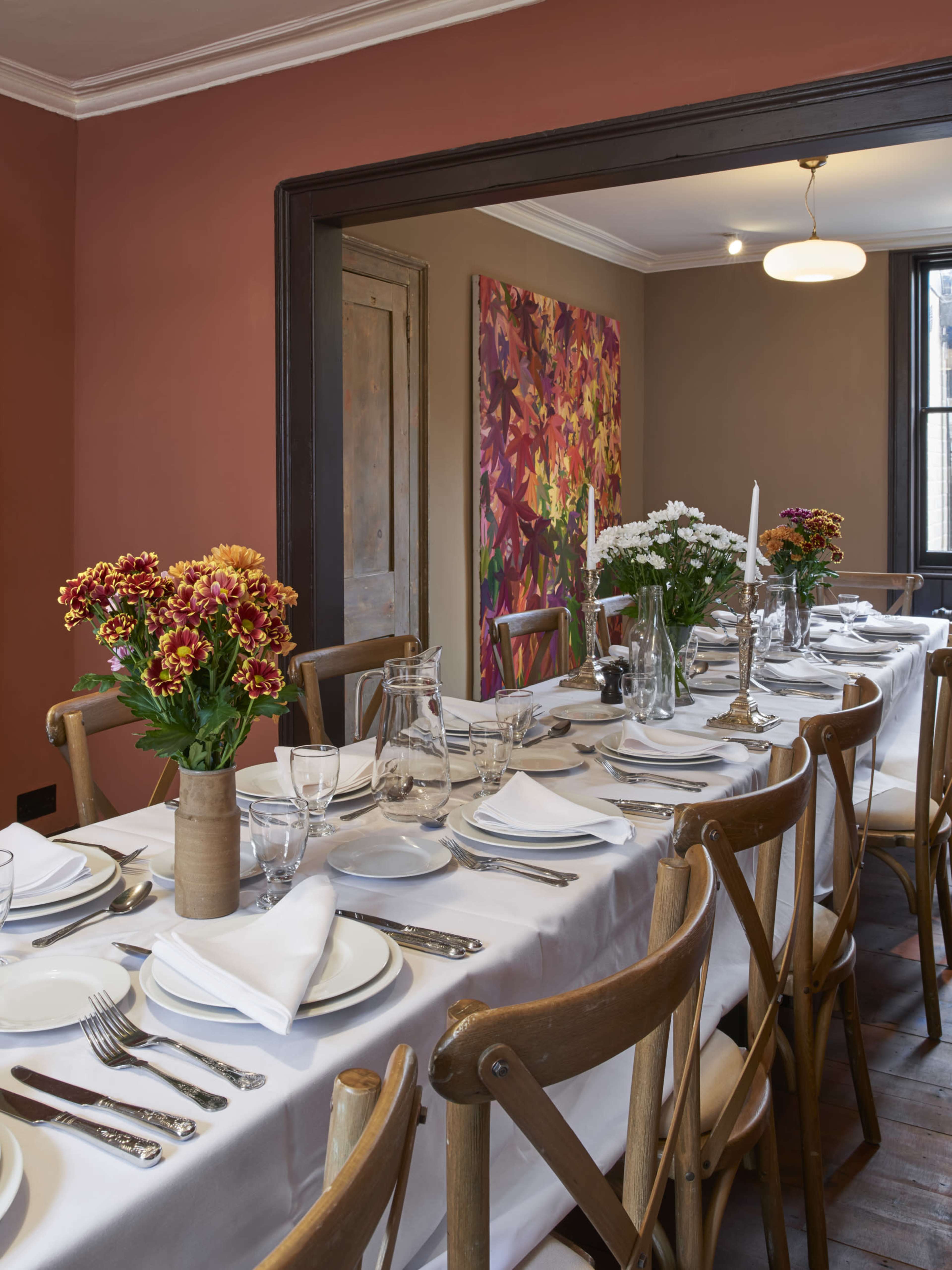 A long dining table set with white tablecloth, ceramic dishes, glassware, and adorned with bouquets of flowers is positioned in a room featuring colorful artwork on the wall.