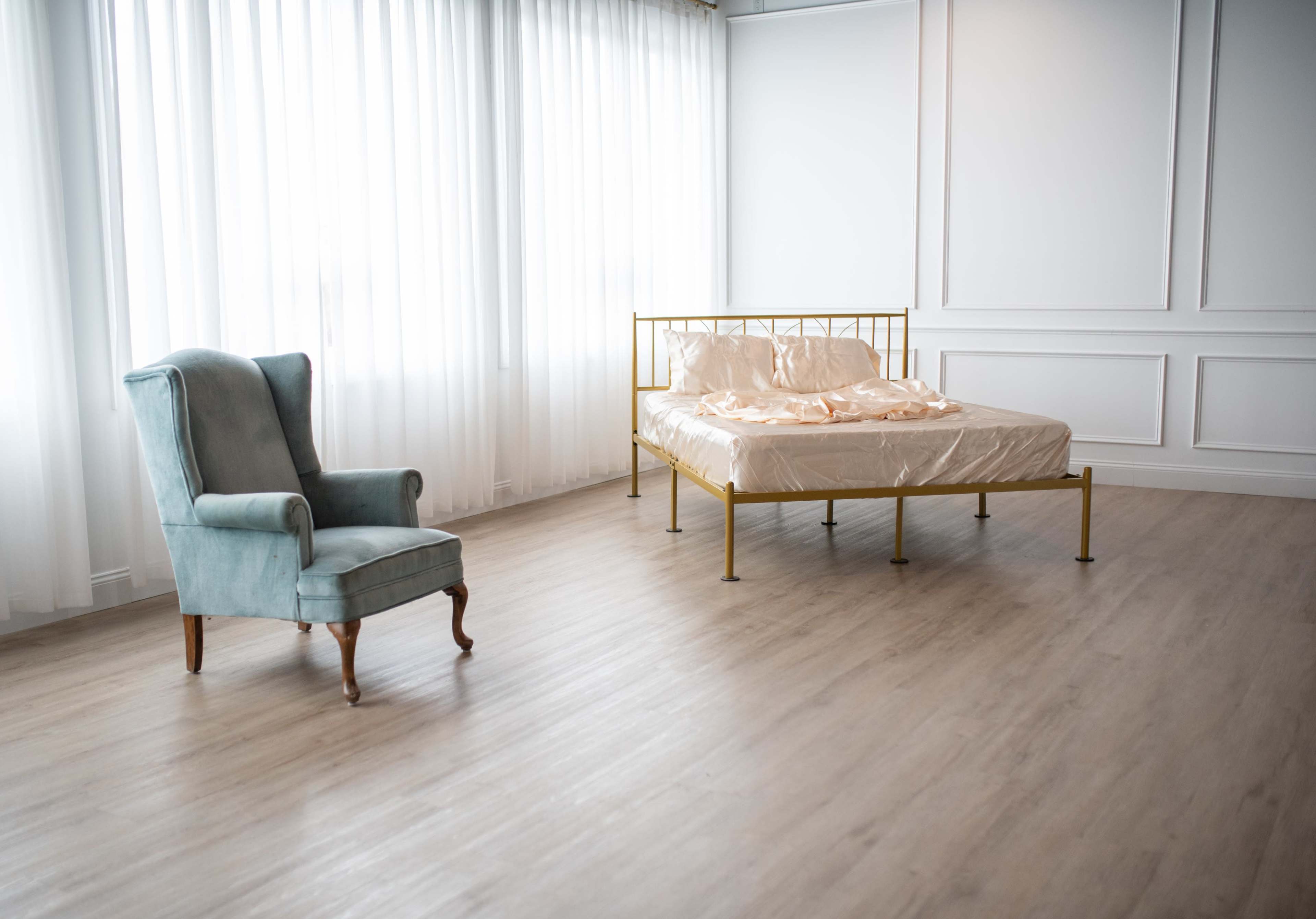 A room features a gold metal bed with unmade sheets and a vintage green upholstered chair, illuminated by sheer white curtains.