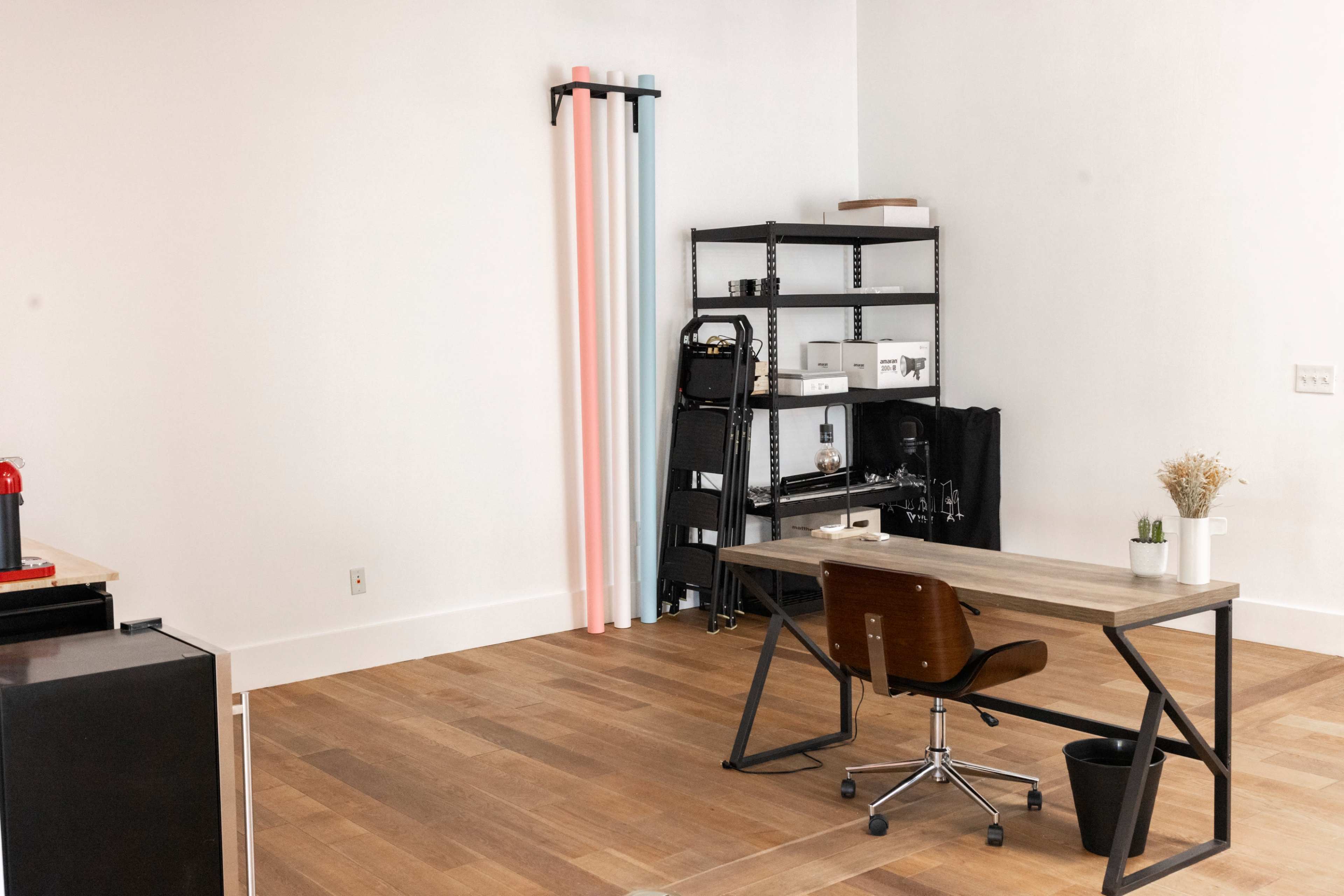 A minimalist office space with a wooden desk, a metal shelving unit, and colorful backdrop rolls against the wall.