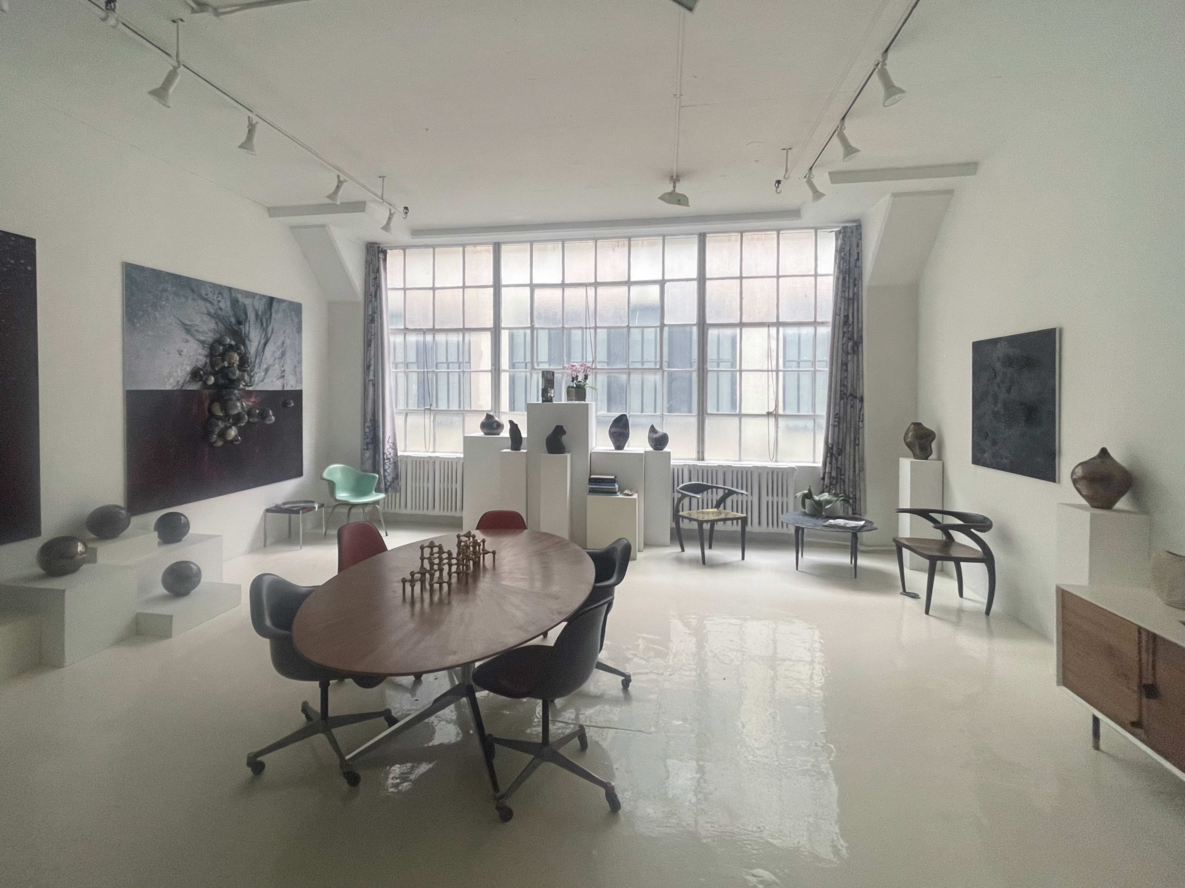 The image shows a spacious, minimalist art studio featuring a large table, several chairs, and various sculptures displayed on pedestals and walls.