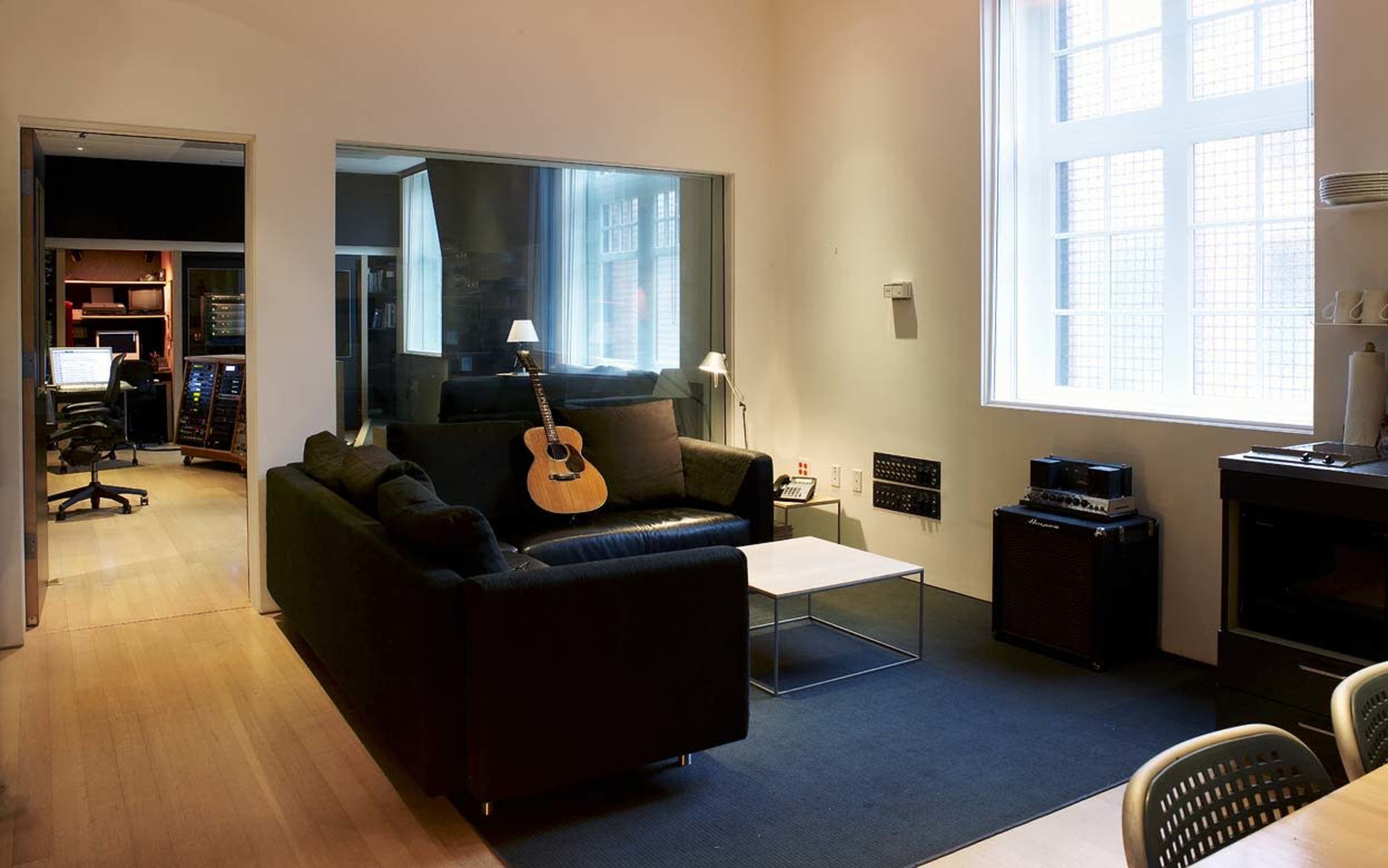 A cozy living room features a black sofa, a coffee table, and a guitar, with large windows providing natural light.