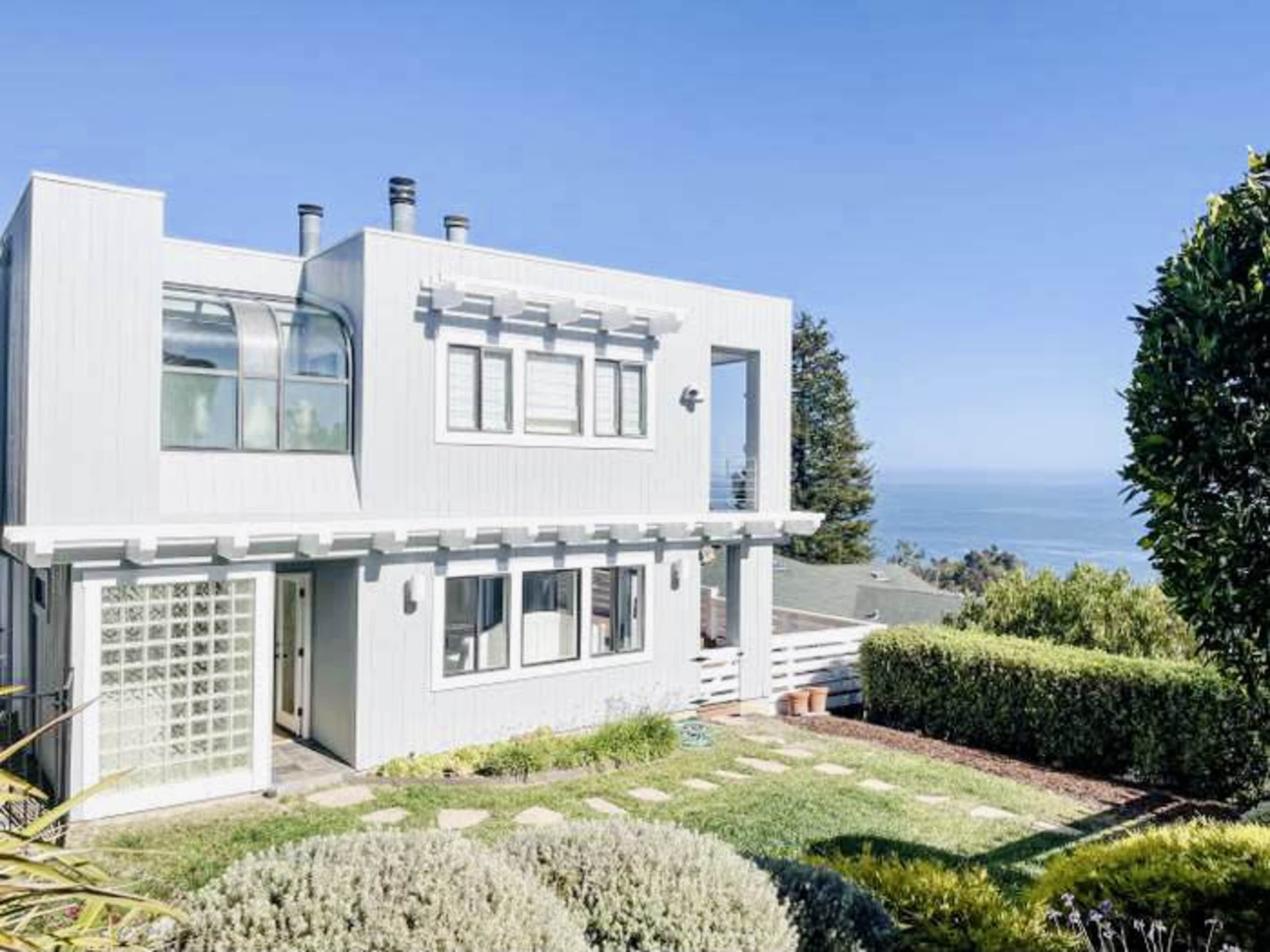 A modern two-story house with a glass block feature and a landscaped yard overlooks a coastal view.