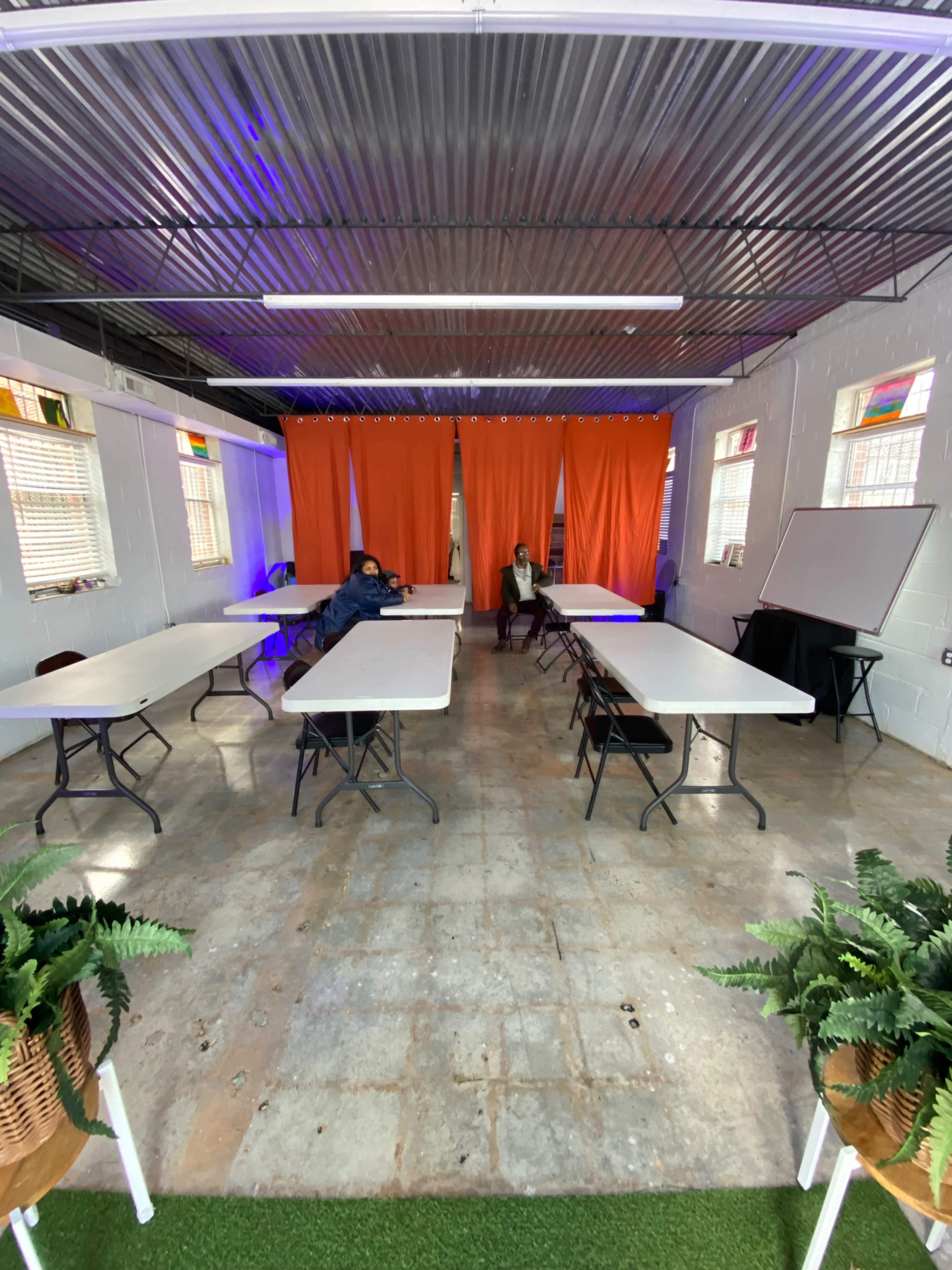 The image shows a sparsely furnished room with several white tables arranged in rows, two people seated at the tables, and orange curtains separating the space.