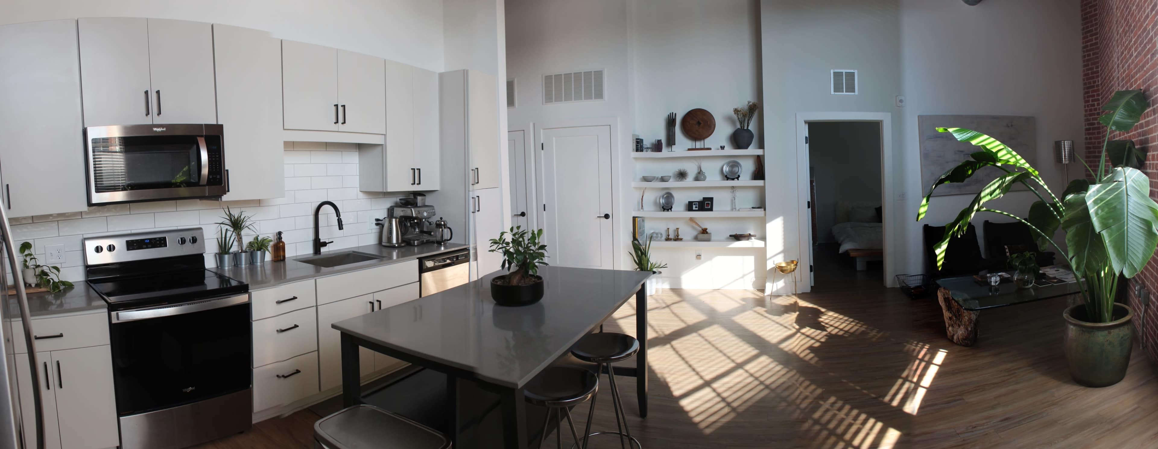 The image shows a modern kitchen with white cabinets and stainless steel appliances, adjacent to a living area featuring shelves with decorative items.