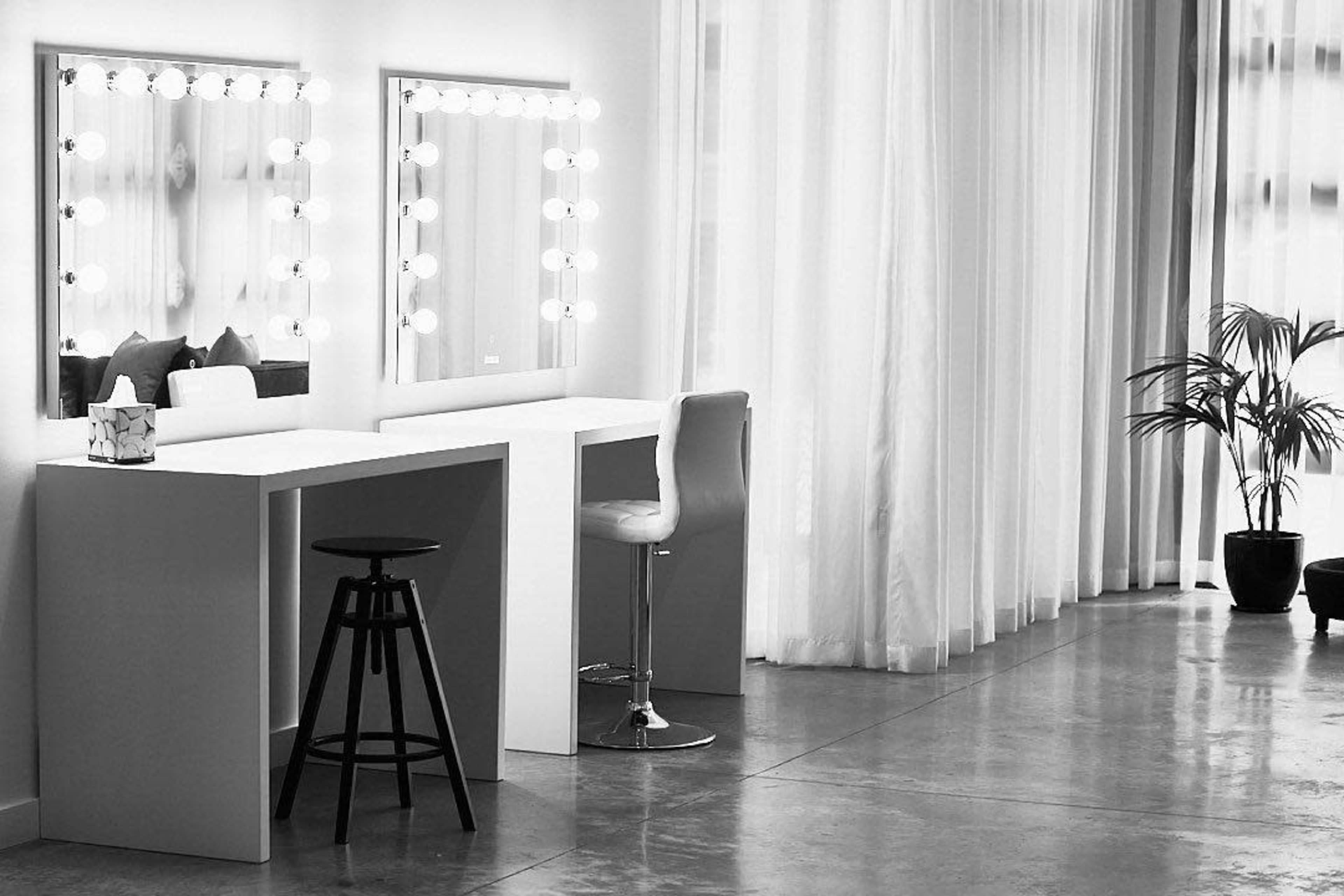 A minimalist makeup room with two lighted mirrors, a bar stool, and a potted plant.