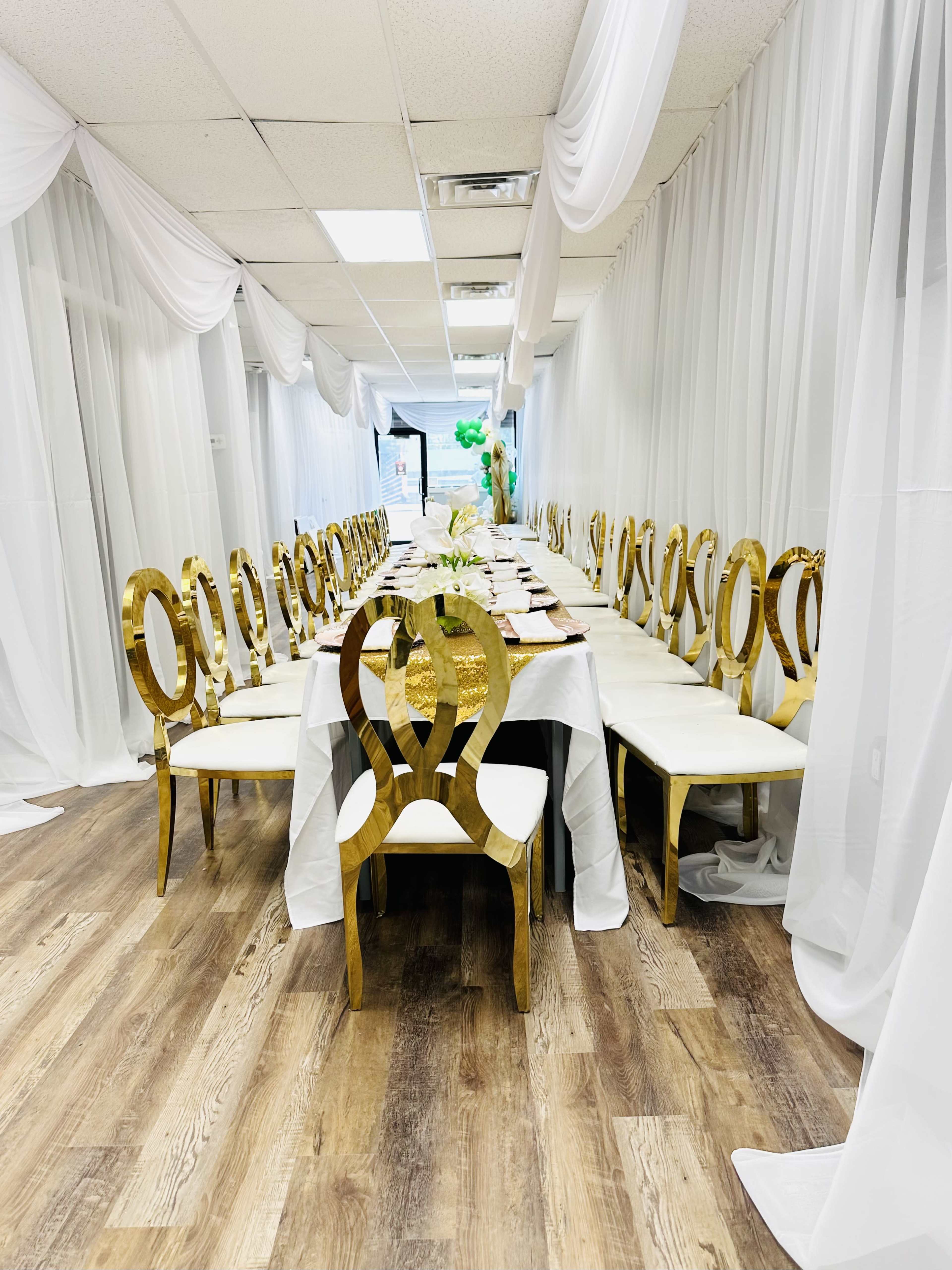 A long dining table is set with white tableware and surrounded by gold chairs, all under draped white fabric in a hallway.