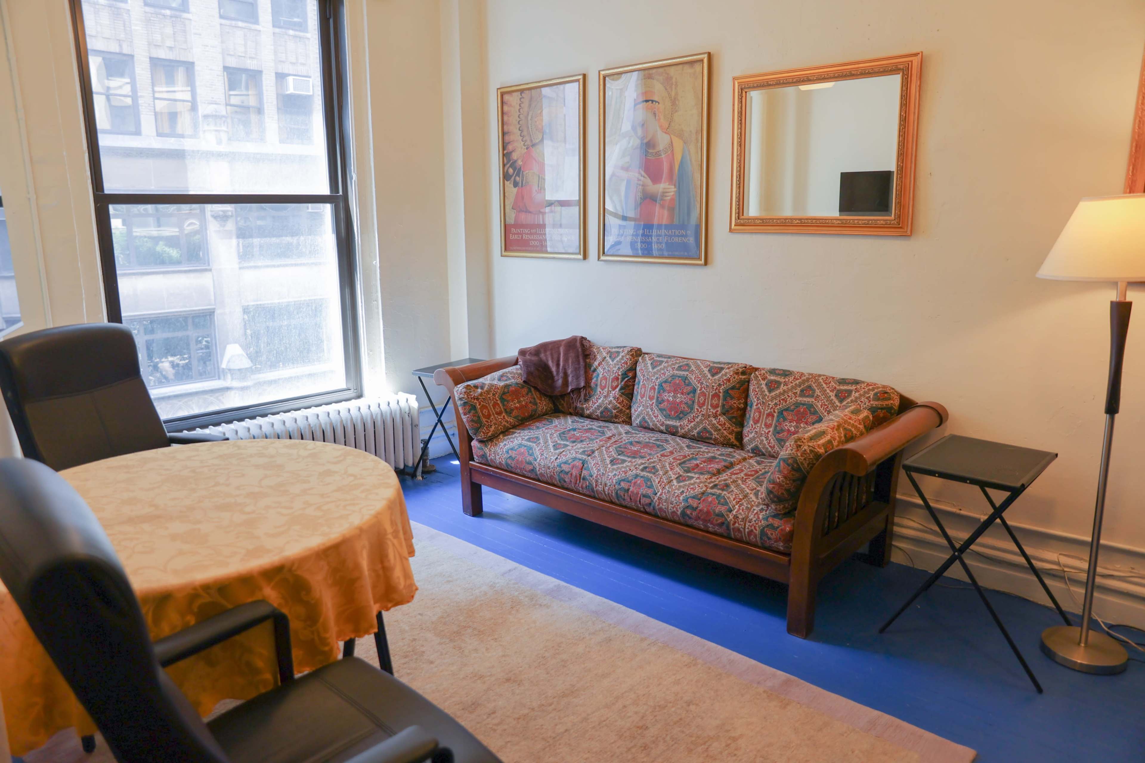 The image shows a living area with a patterned sofa, a round table with a tablecloth, two black chairs, and framed artwork on the walls.