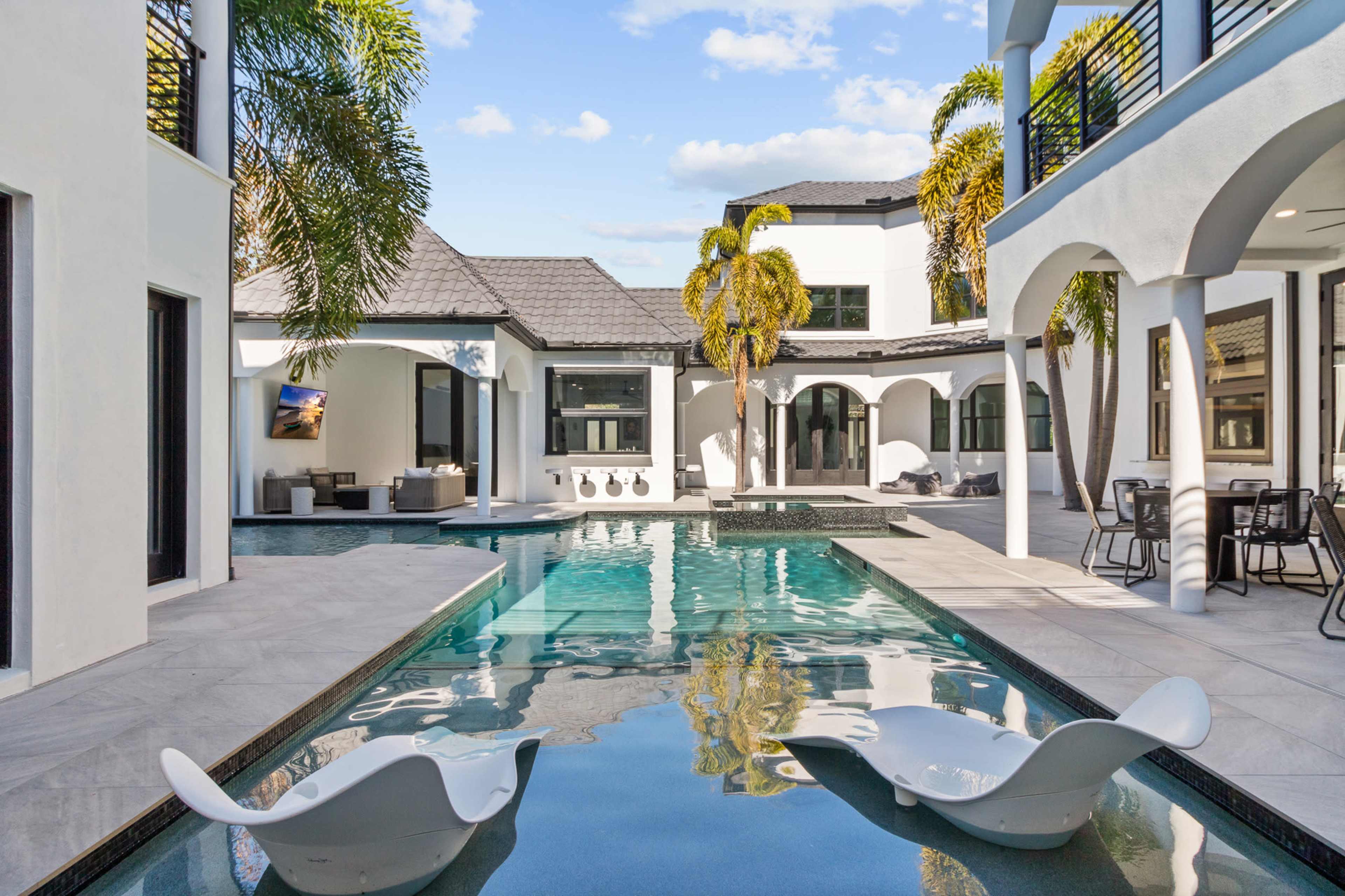 The image shows a modern villa with a swimming pool featuring white lounge chairs and palm trees, surrounded by architectural elements of the house.
