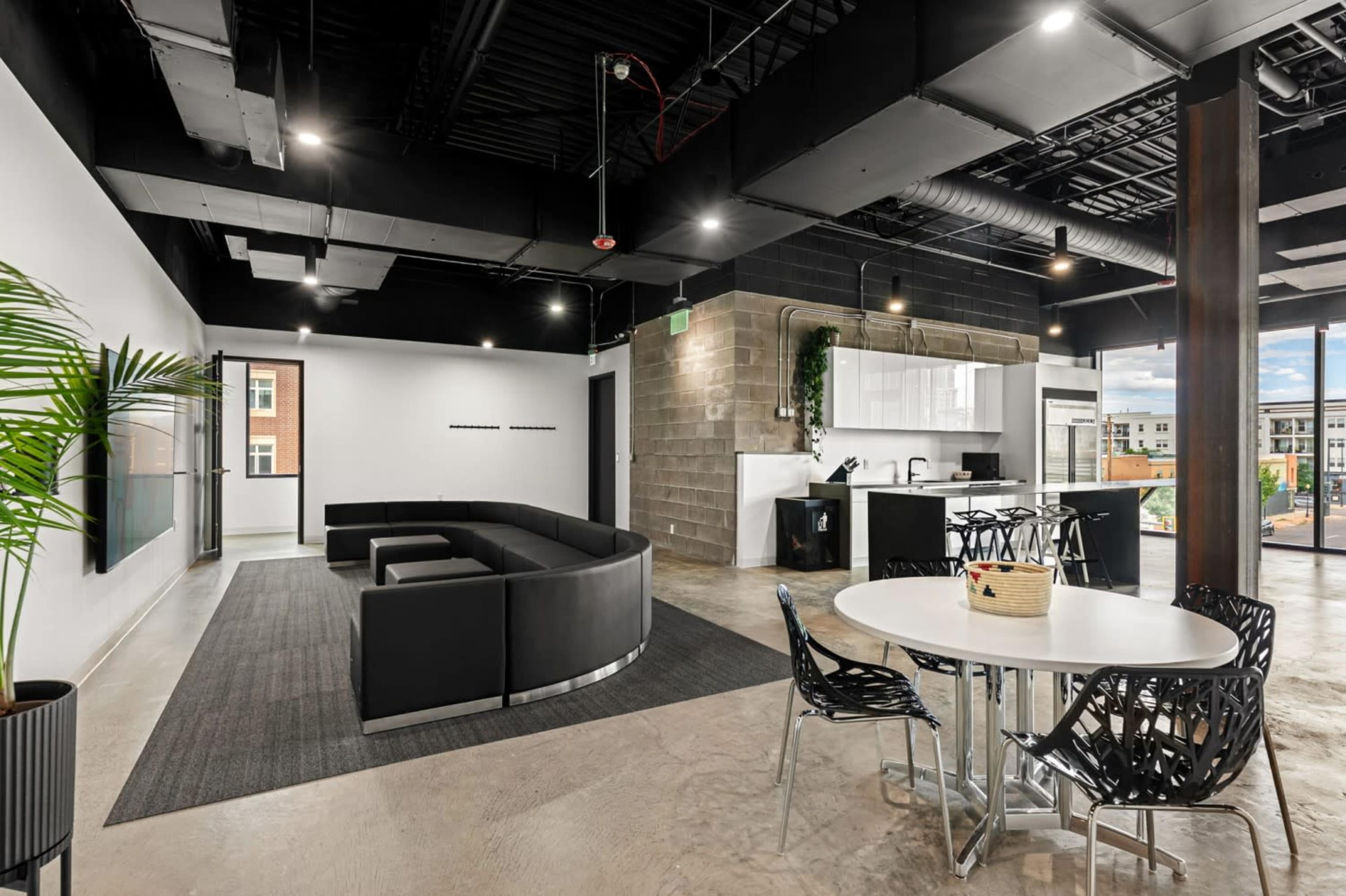 The image shows a modern open office space with a black curved sofa, a round dining table, and a kitchen area in the background.