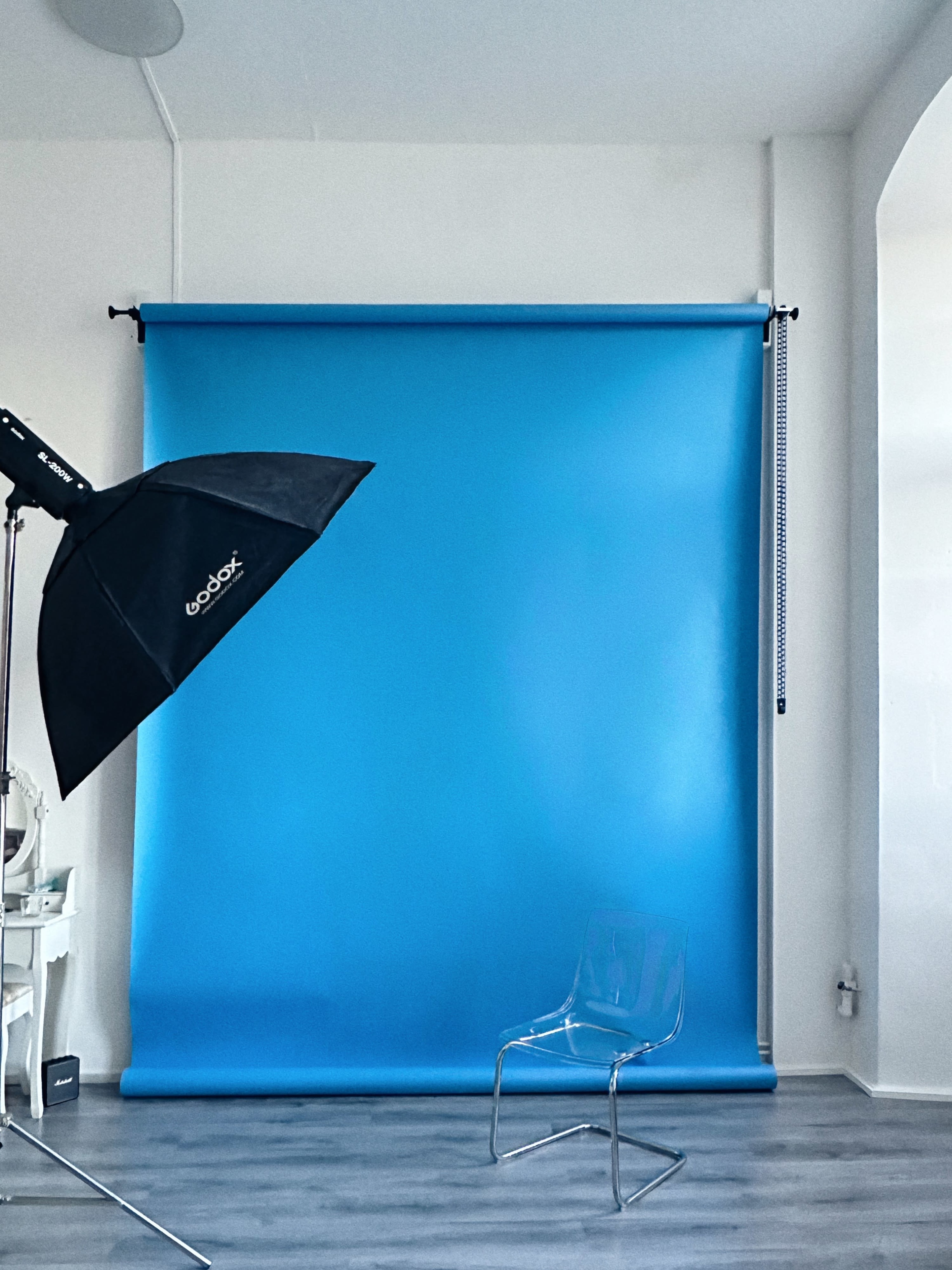 A photography studio with a blue backdrop, an umbrella softbox light, and a transparent chair on a wooden floor.