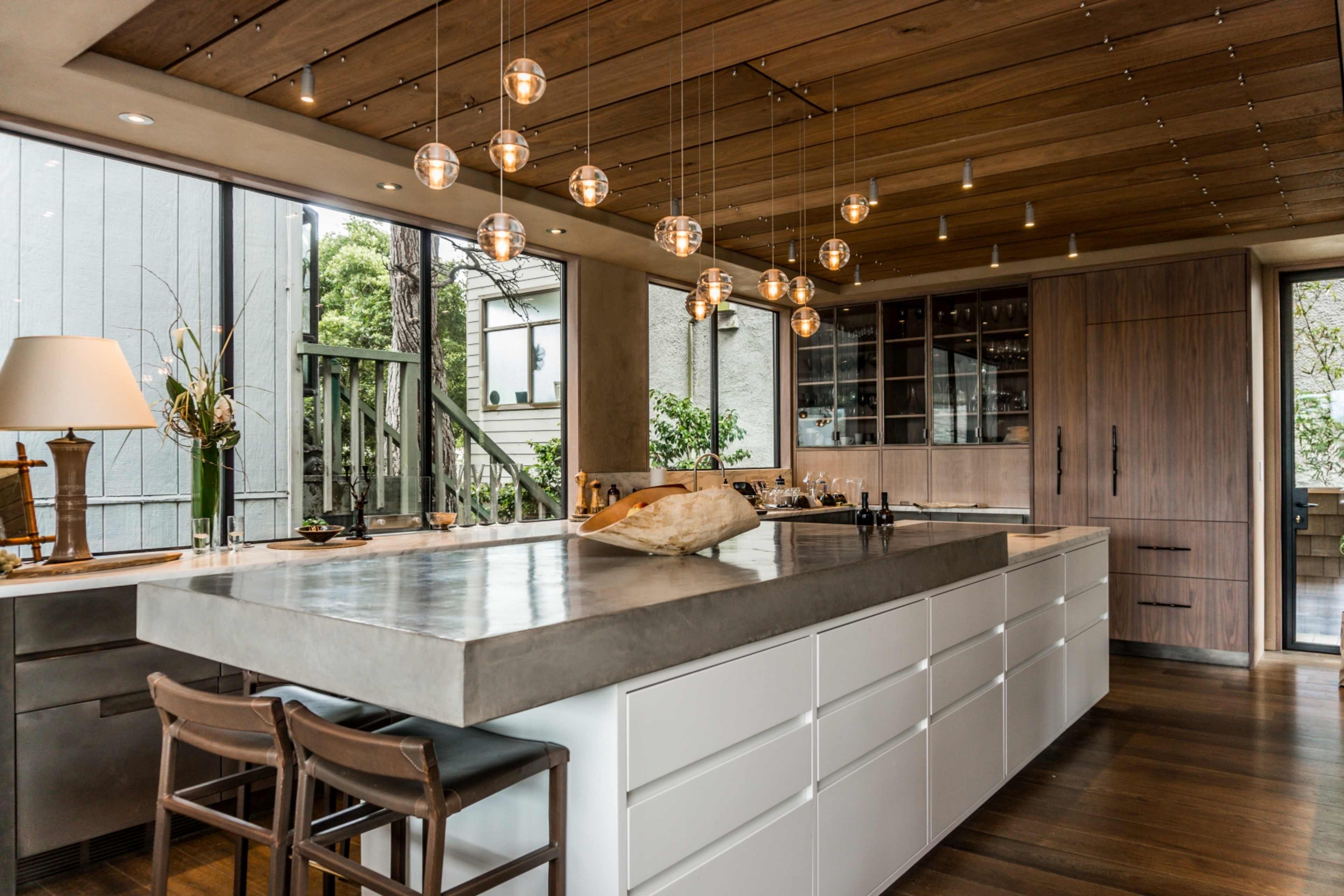 The kitchen features a large concrete island with wooden cabinetry, modern pendant lighting, and large windows that provide natural light.