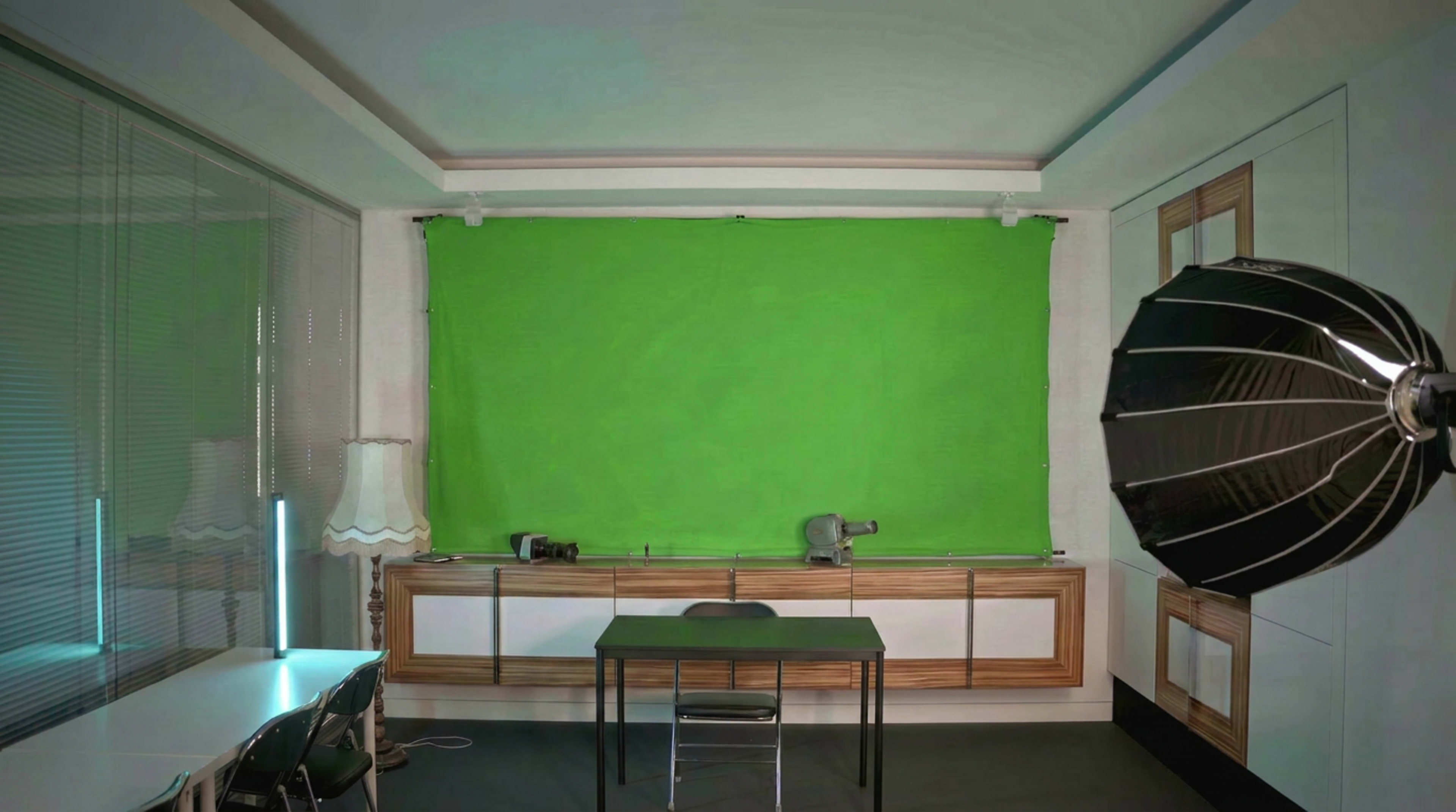The image shows a room with a green screen backdrop, a black chair, a table, and studio lighting equipment on one side.