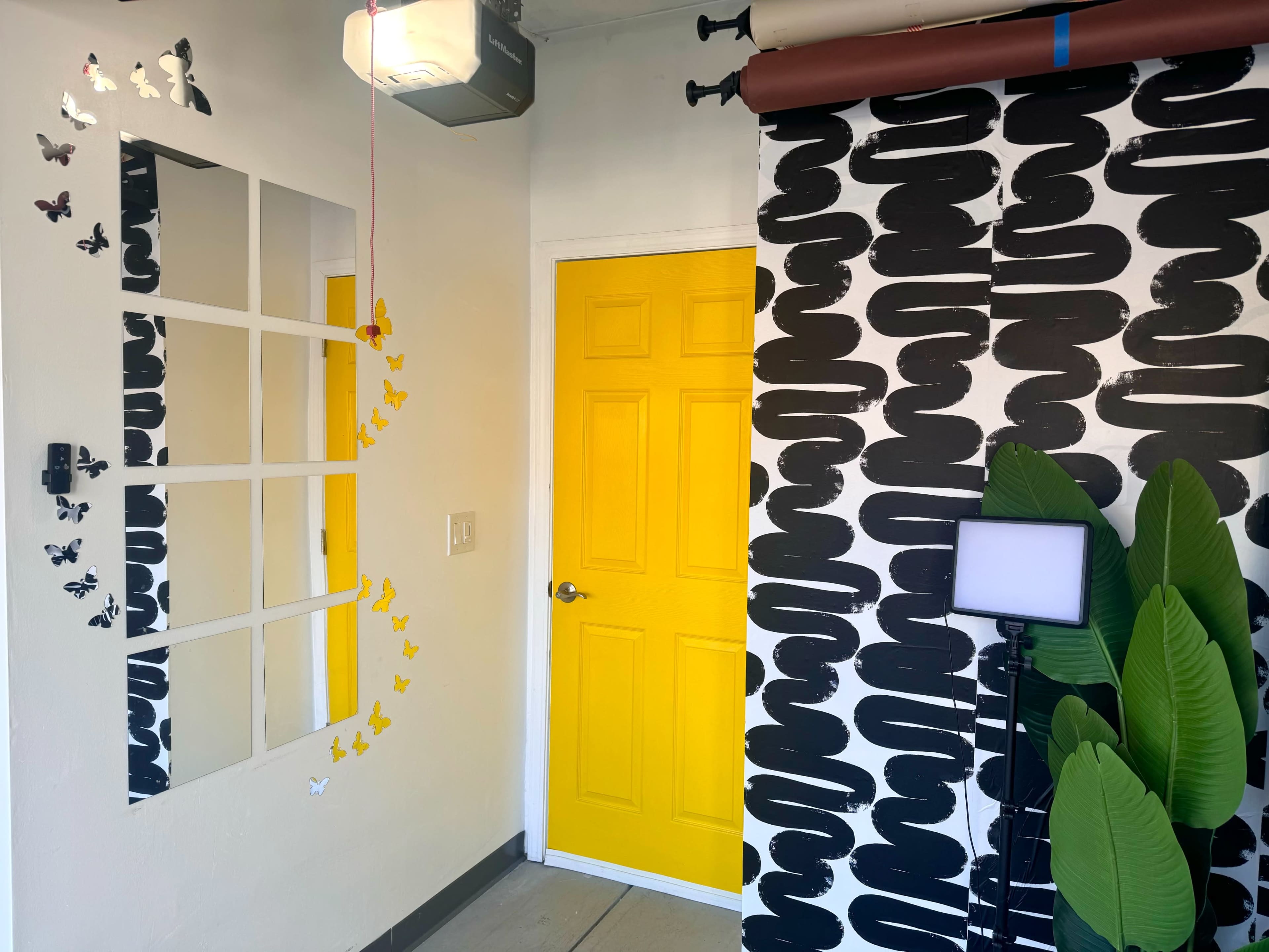 The image shows a brightly lit corner of a room featuring a yellow door, a mirror with a grid pattern, a patterned wall, and decorative butterflies alongside a floor lamp and large green leaves.