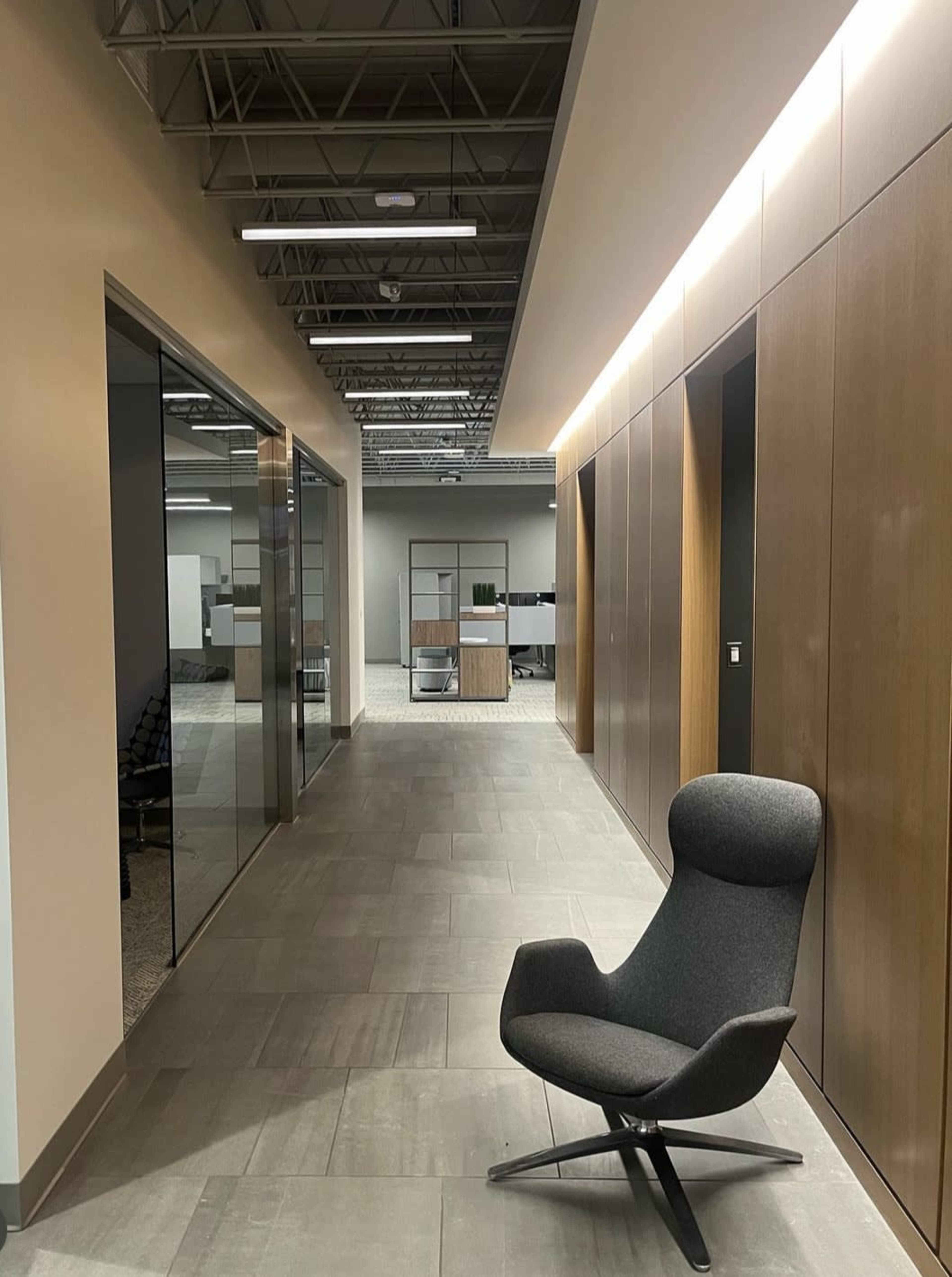 A modern office hallway with a gray chair positioned next to glass partitioned rooms and wooden paneling on the walls.