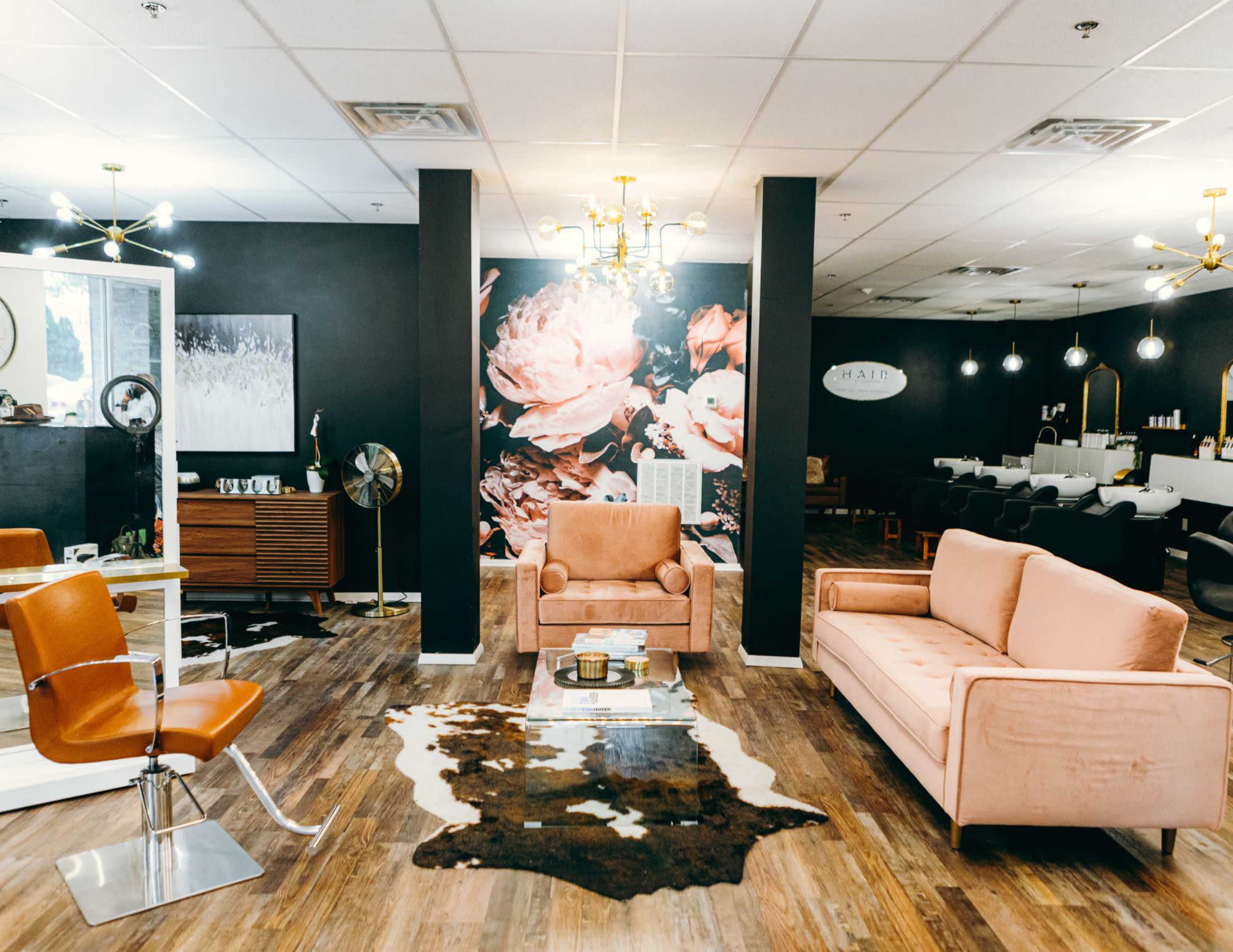 A modern salon features a pink velvet sofa, a cowhide rug, and a wall adorned with floral artwork, set against dark walls and bright lighting.