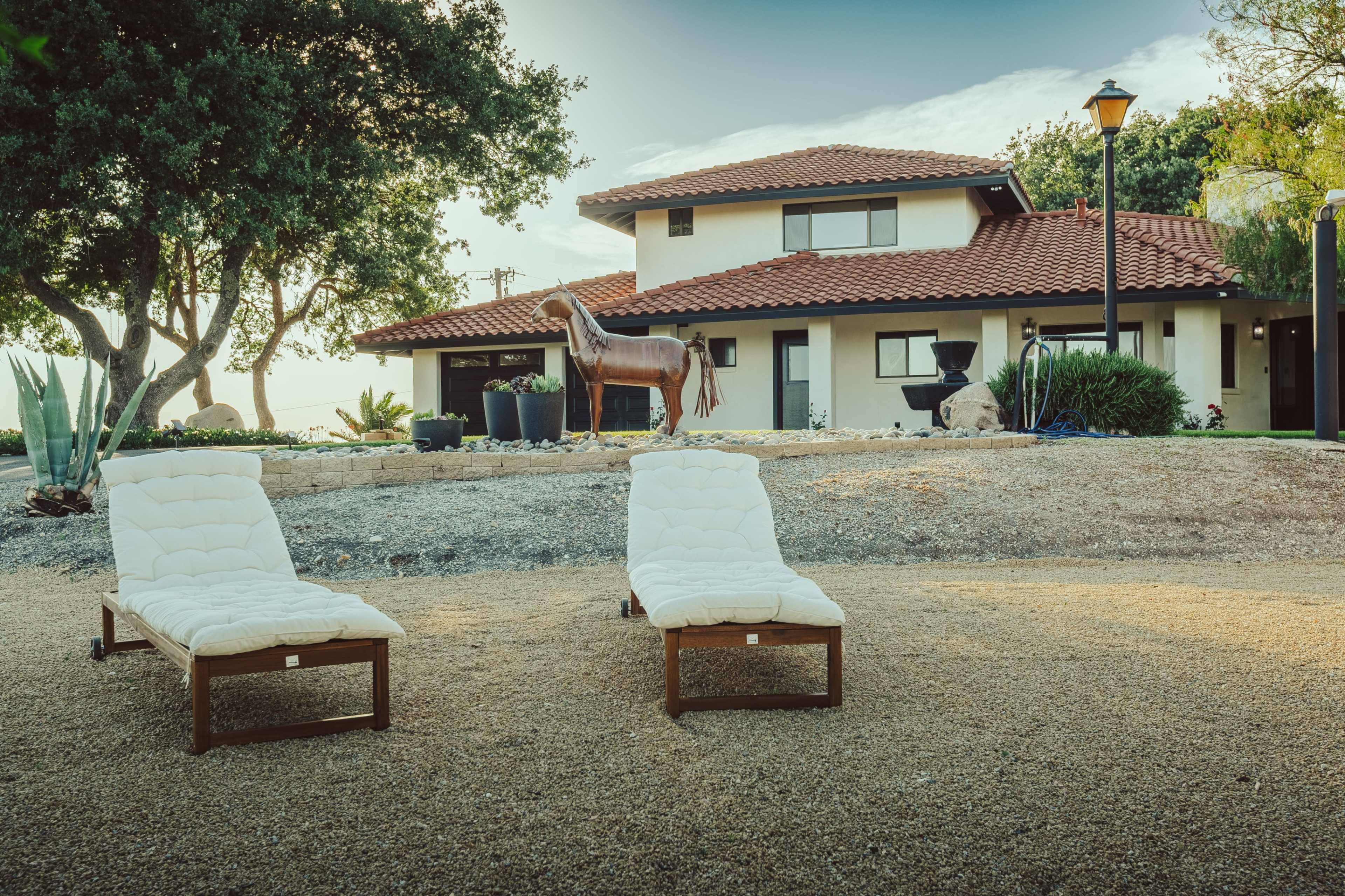 The image features two white lounge chairs on a gravel area in front of a modern, two-story house with a red tile roof and a horse sculpture in the yard.