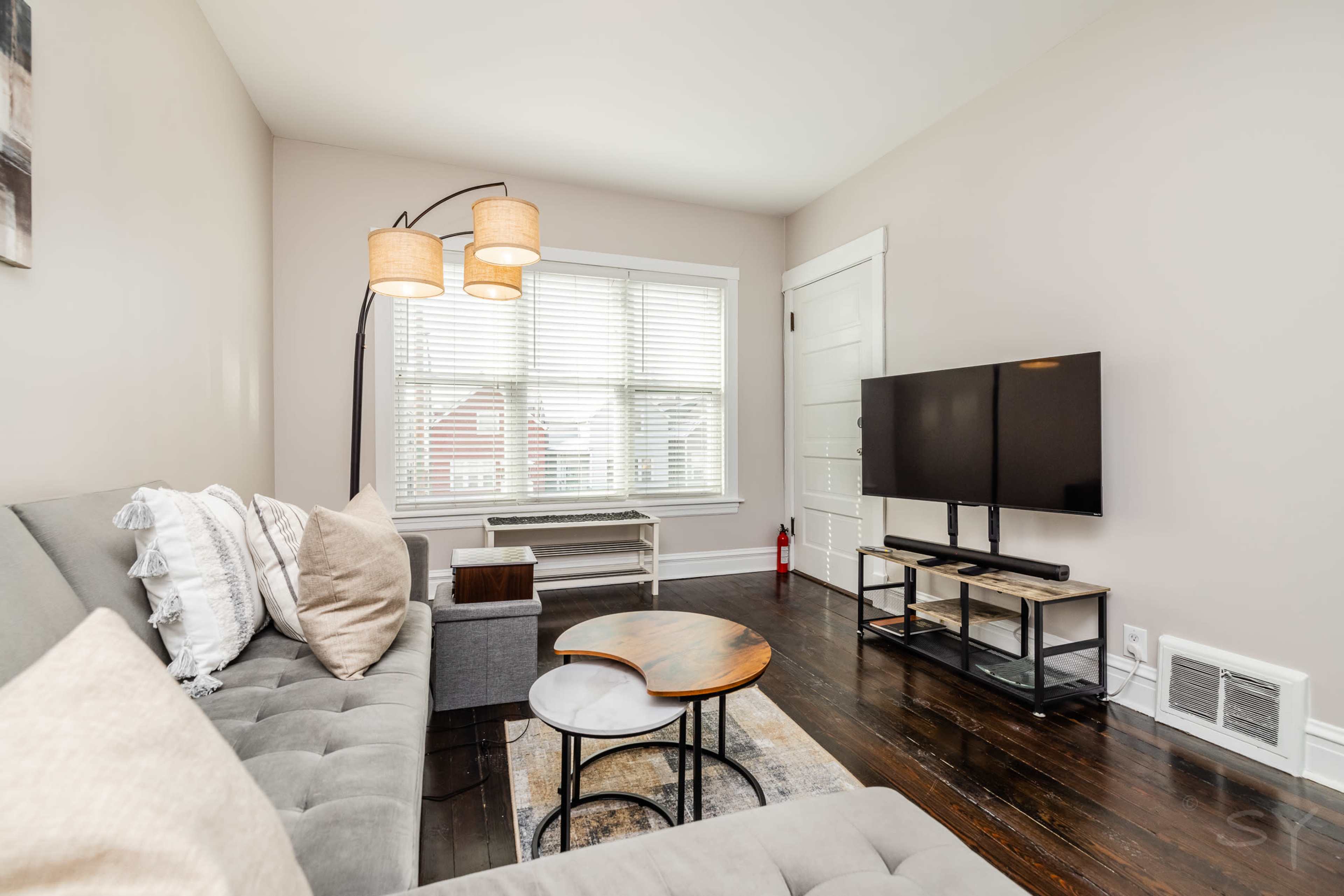 A living room with a gray sofa, a wooden coffee table, a floor lamp with two shades, a television on a stand, and a window with white blinds.