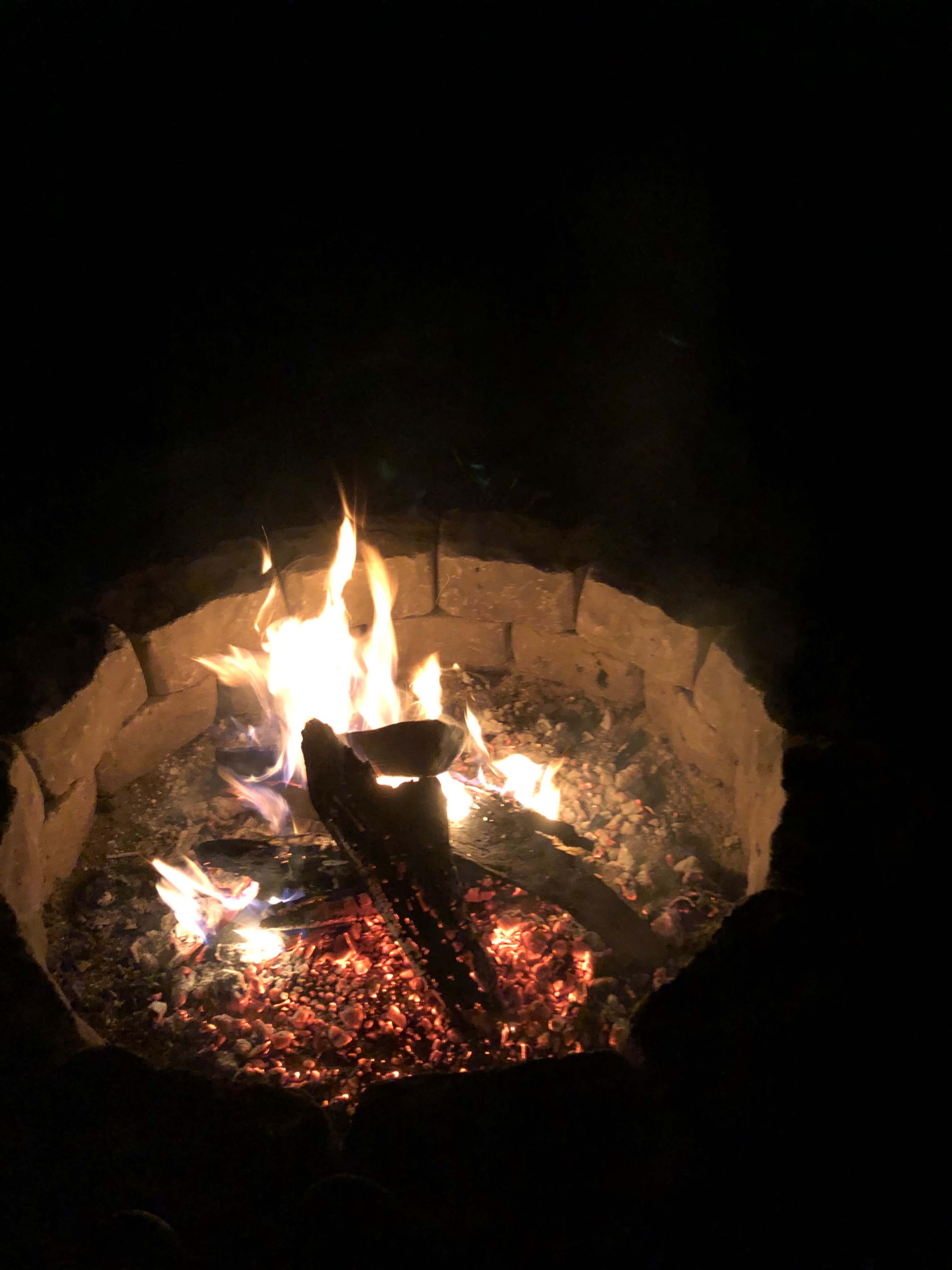 A fire burns in a stone fire pit surrounded by dark surroundings.