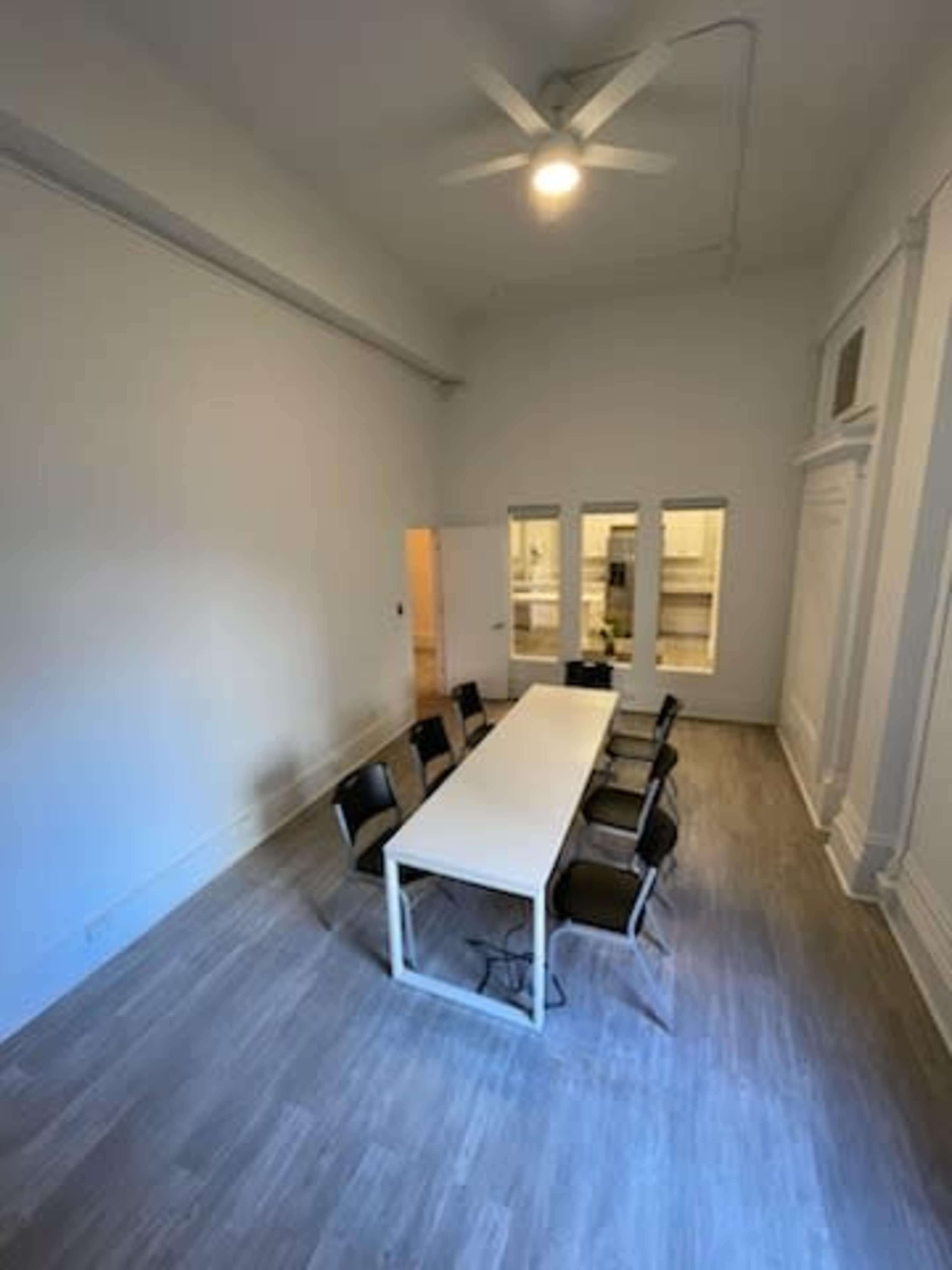 The image shows a spacious meeting room with a long white table and several chairs, featuring large windows and a ceiling fan.