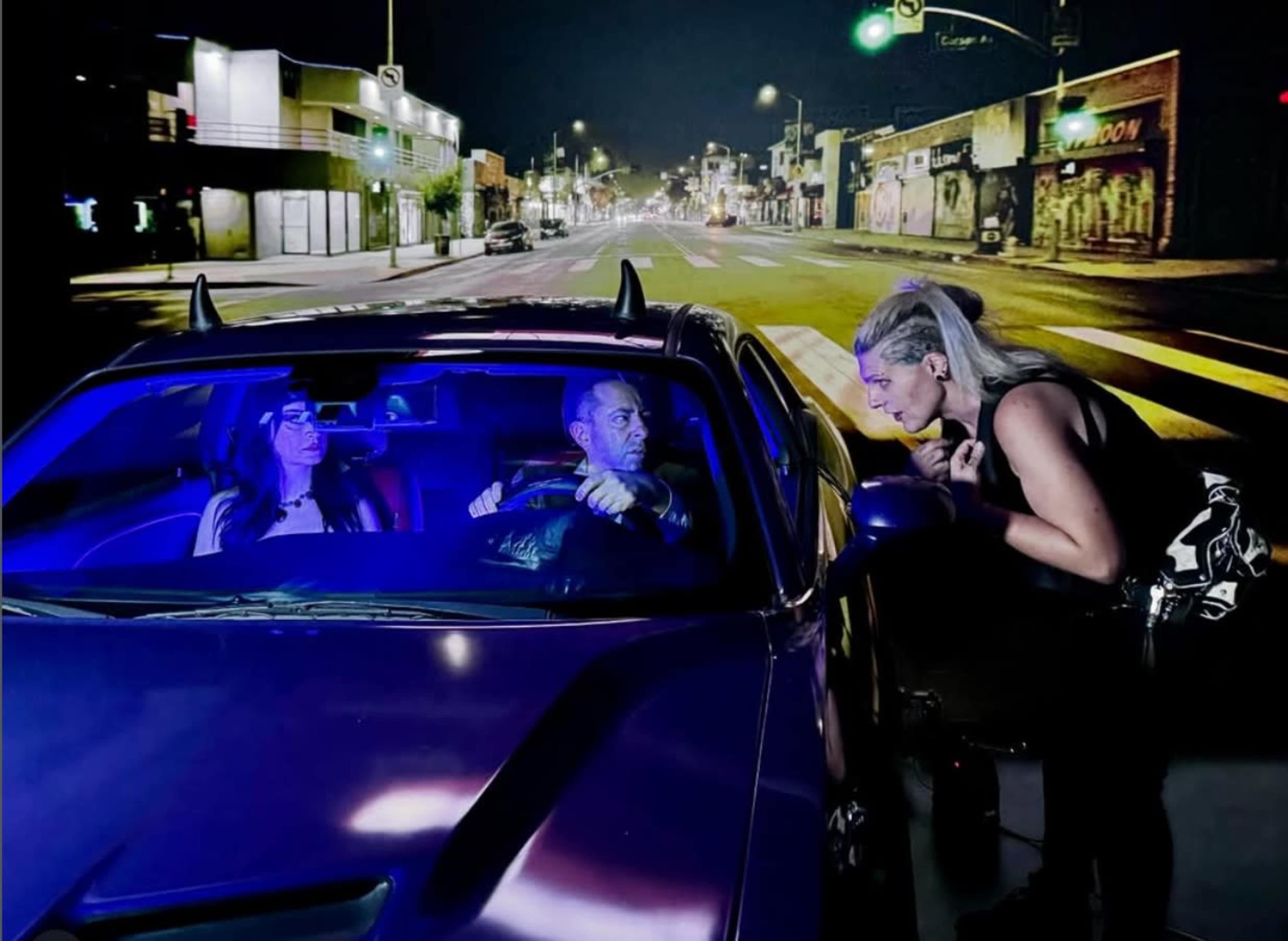A man and a woman sit in a car at night while another woman leans toward the driver's side, illuminated by blue and purple lights.