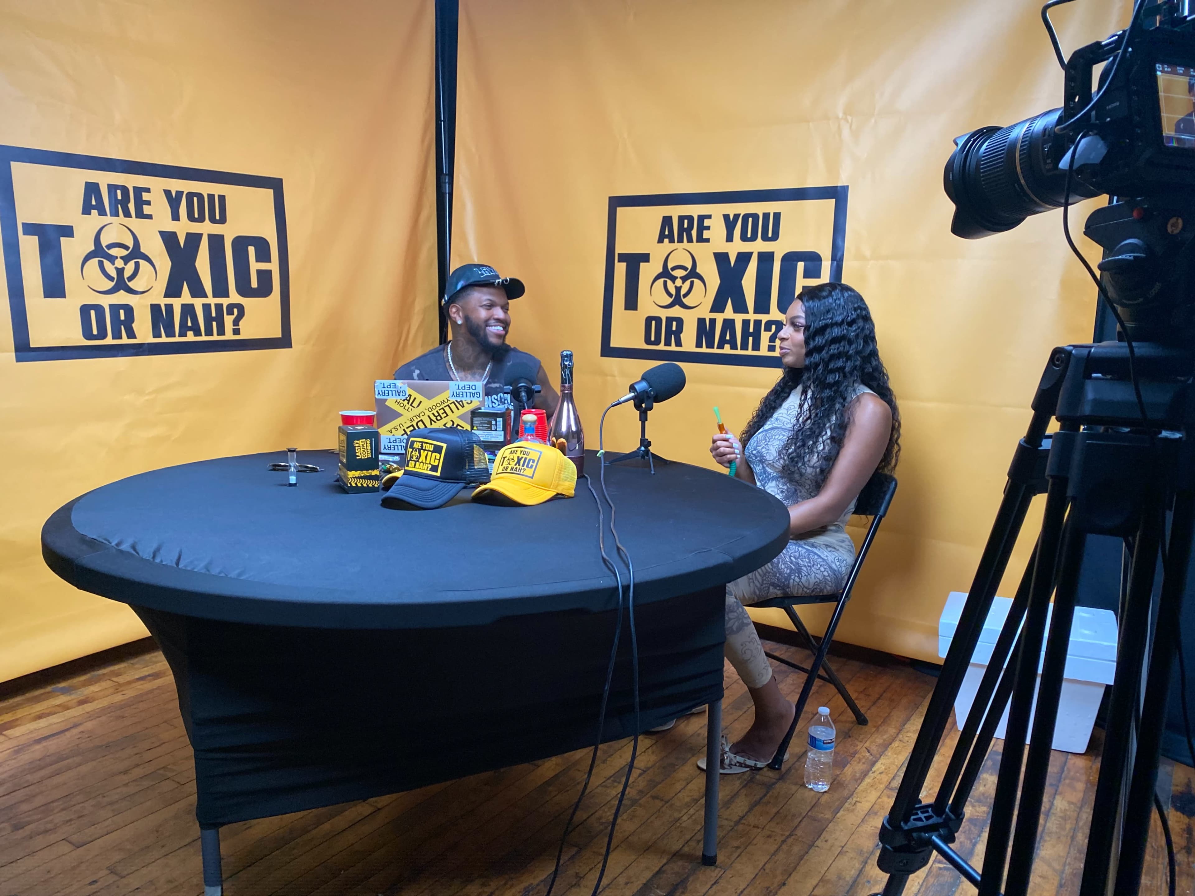 A man and a woman sit at a round table with branded merchandise in front of a backdrop that reads "ARE YOU TOXIC OR NAH?"