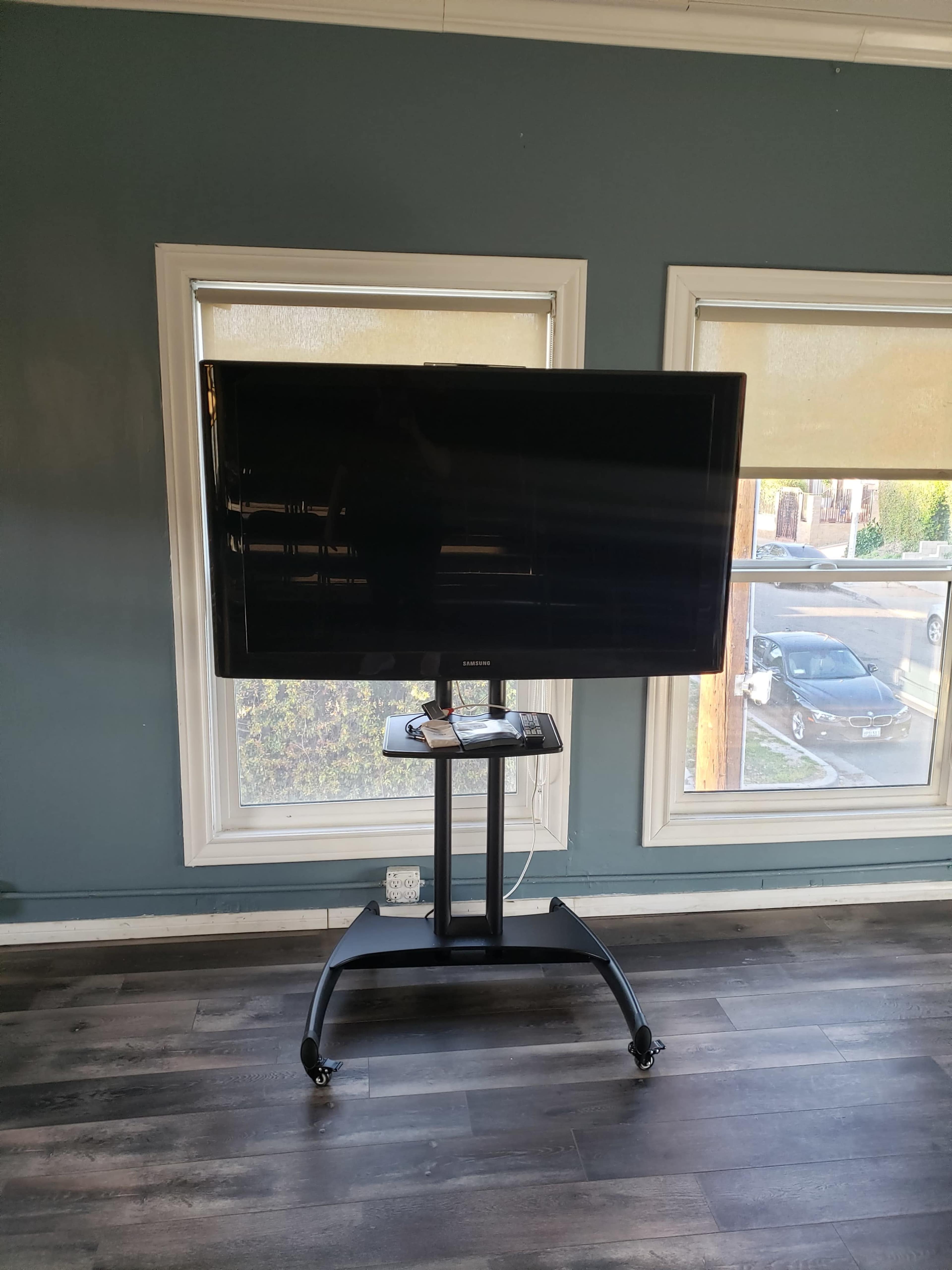 A large, black flat-screen TV is mounted on a wheeled stand in a room with two windows and a wooden floor.