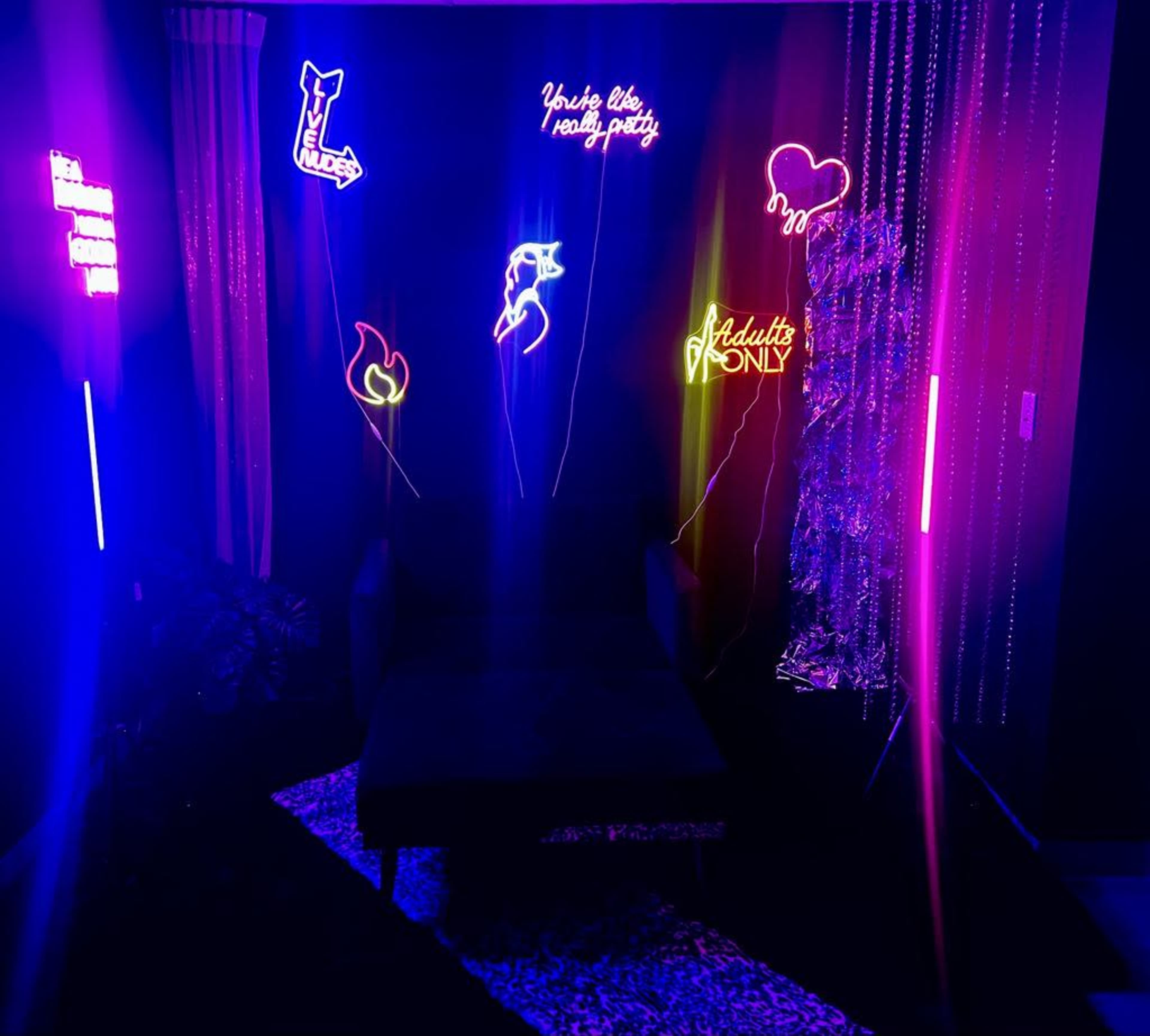 The image shows a dark room with a black couch, illuminated by various colorful neon signs and lights, including phrases like "You're like really pretty" and "Adults Only."